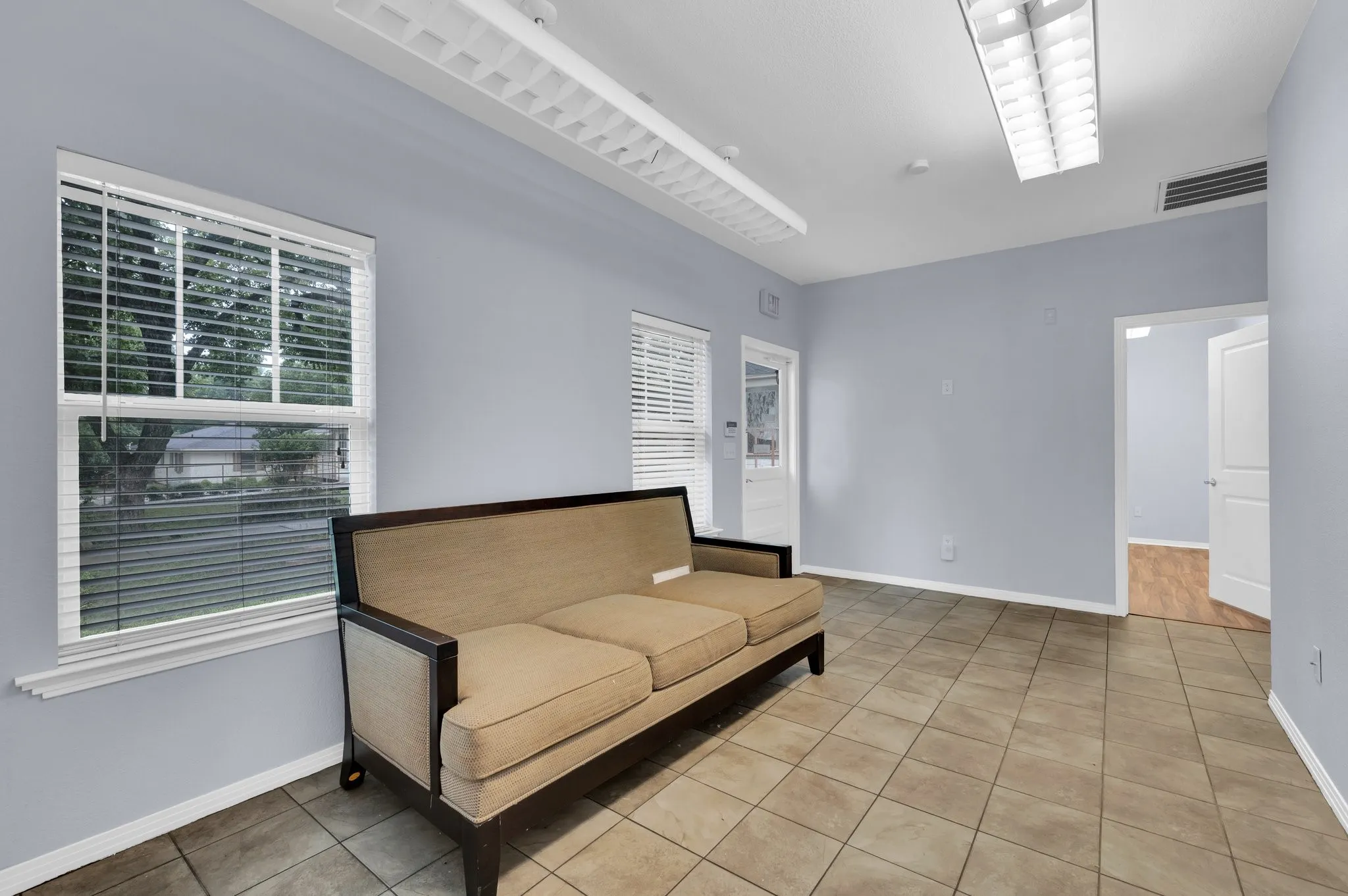 Spacious room featuring neutral tile flooring, light blue walls, and two large windows with blinds