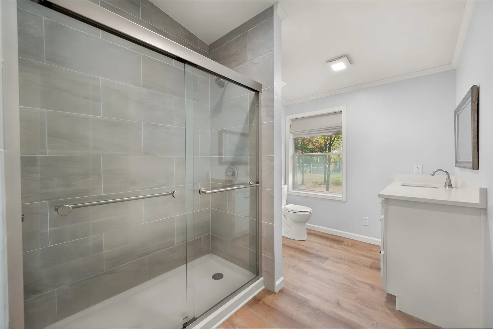 Glass-enclosed shower with tile surround, single vanity with integrated sink, wood-finish flooring, and a window with a shade
