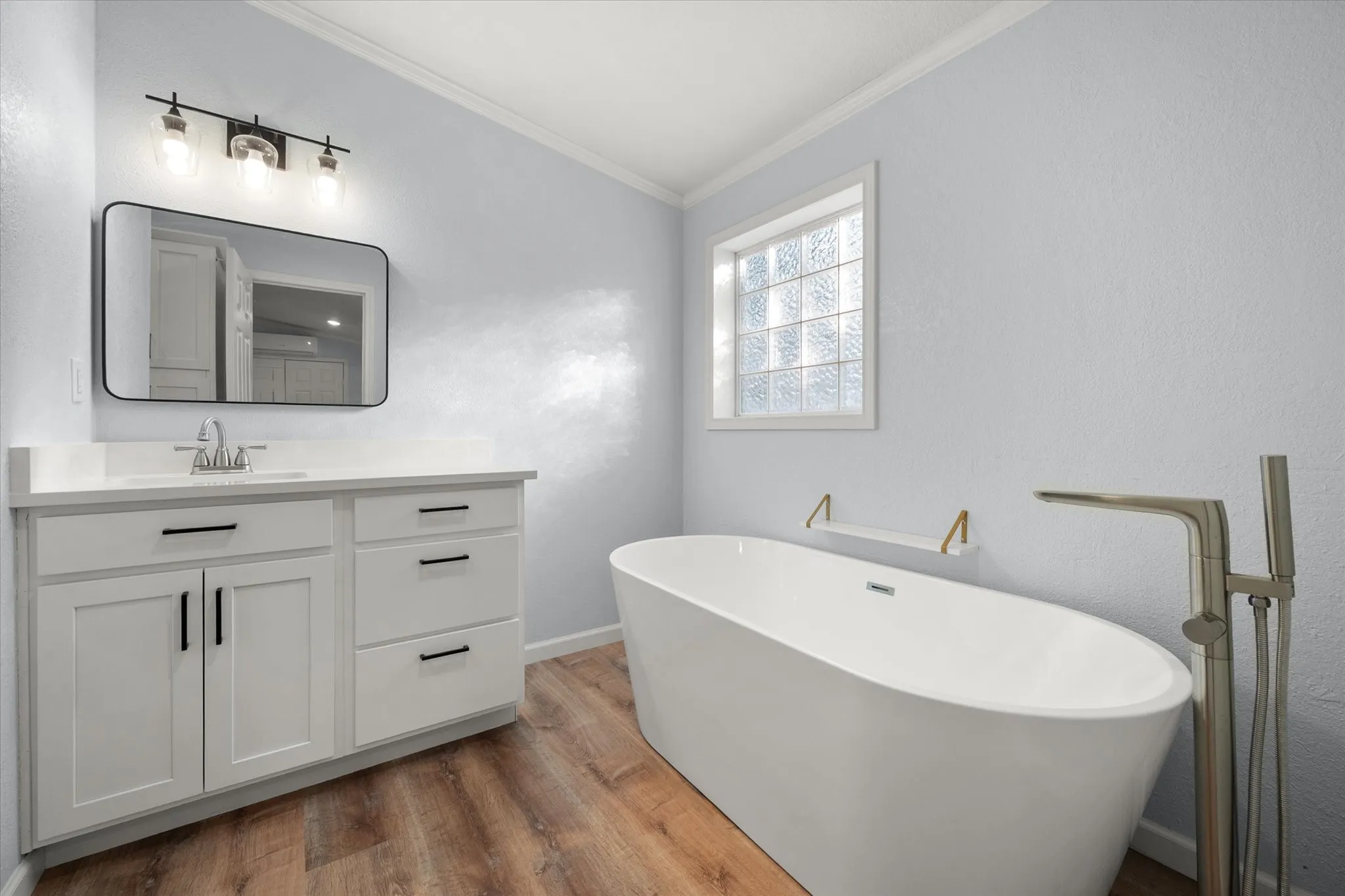 Freestanding soaking tub with floor-mounted faucet, wood-finish flooring, white vanity with black hardware, rectangular vanity mirror, and window with privacy glass blocks