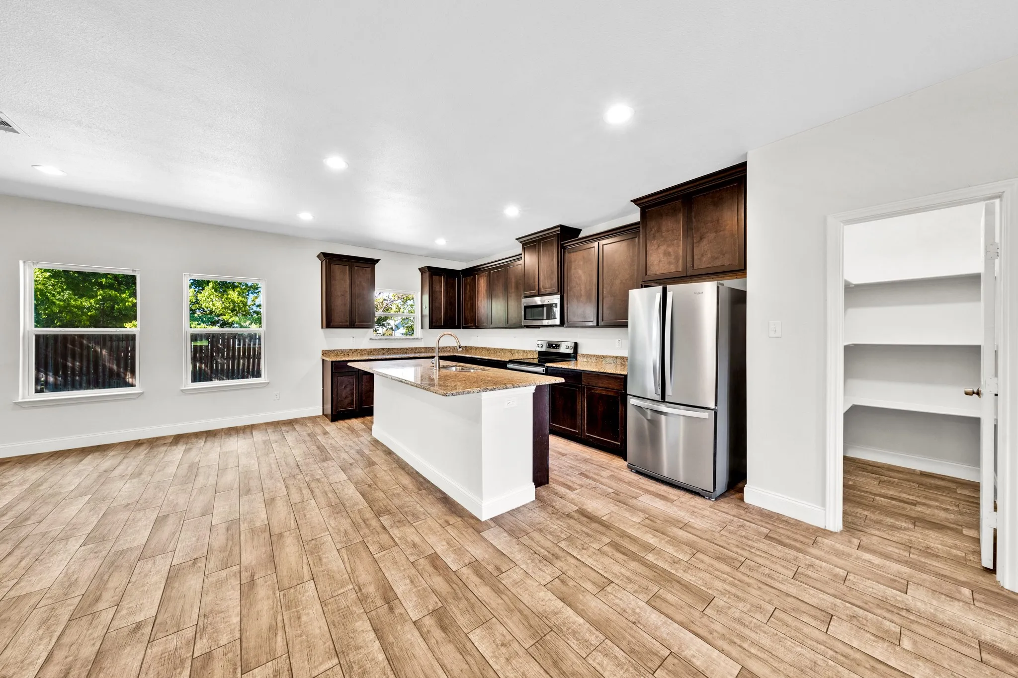 Open-concept kitchen with wood-finish flooring, dark wood cabinetry, stainless steel appliances, a center island with sink, and a walk-in pantry