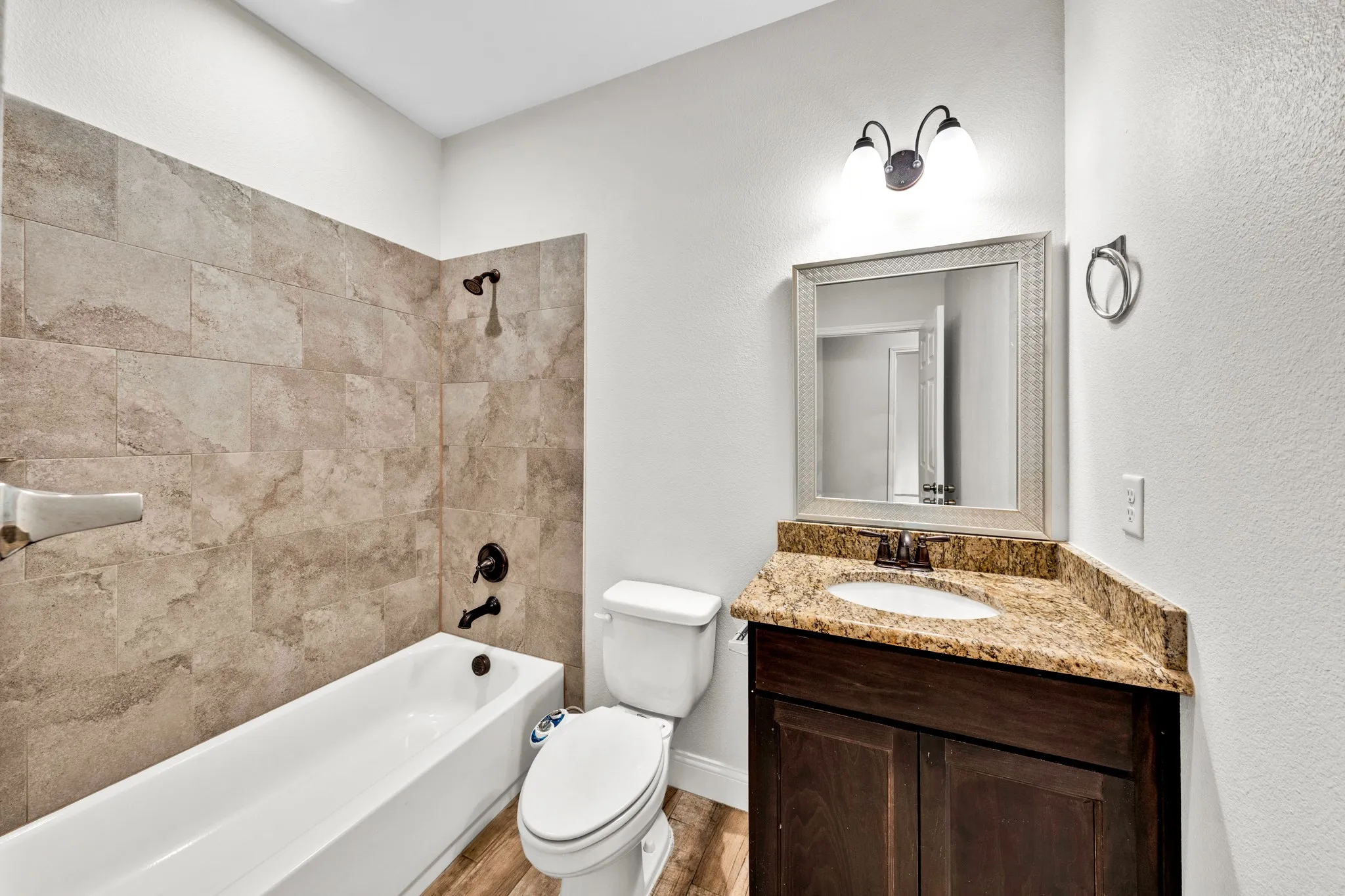 Bathroom featuring a white tub, natural stone-look tile shower surround, wood-finish flooring, dark wood vanity with a light-toned stone countertop, and a framed mirror