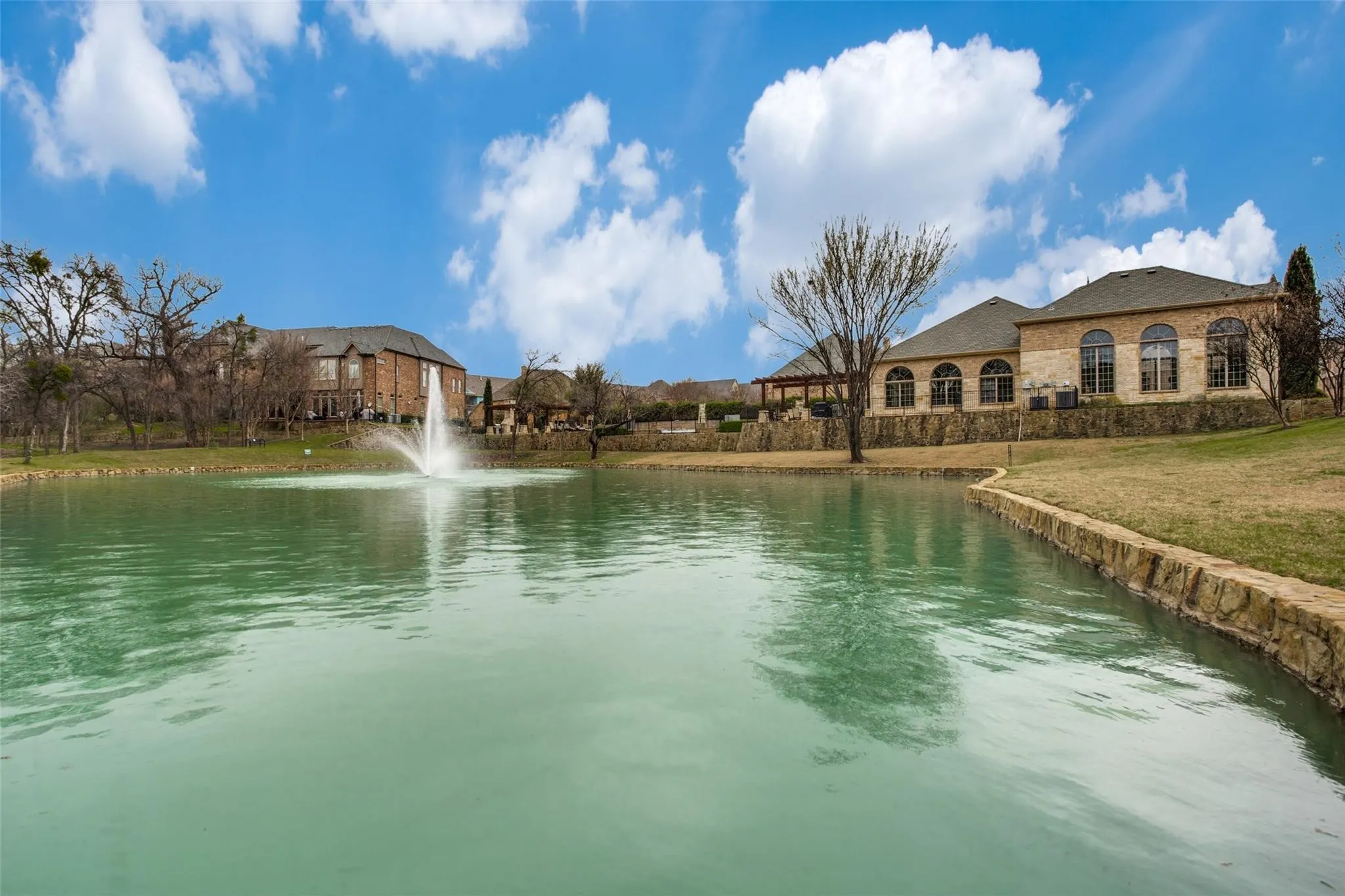 Expansive waterfront property featuring a stone-faced exterior, arched windows, covered outdoor living space, and a private pond with a fountain