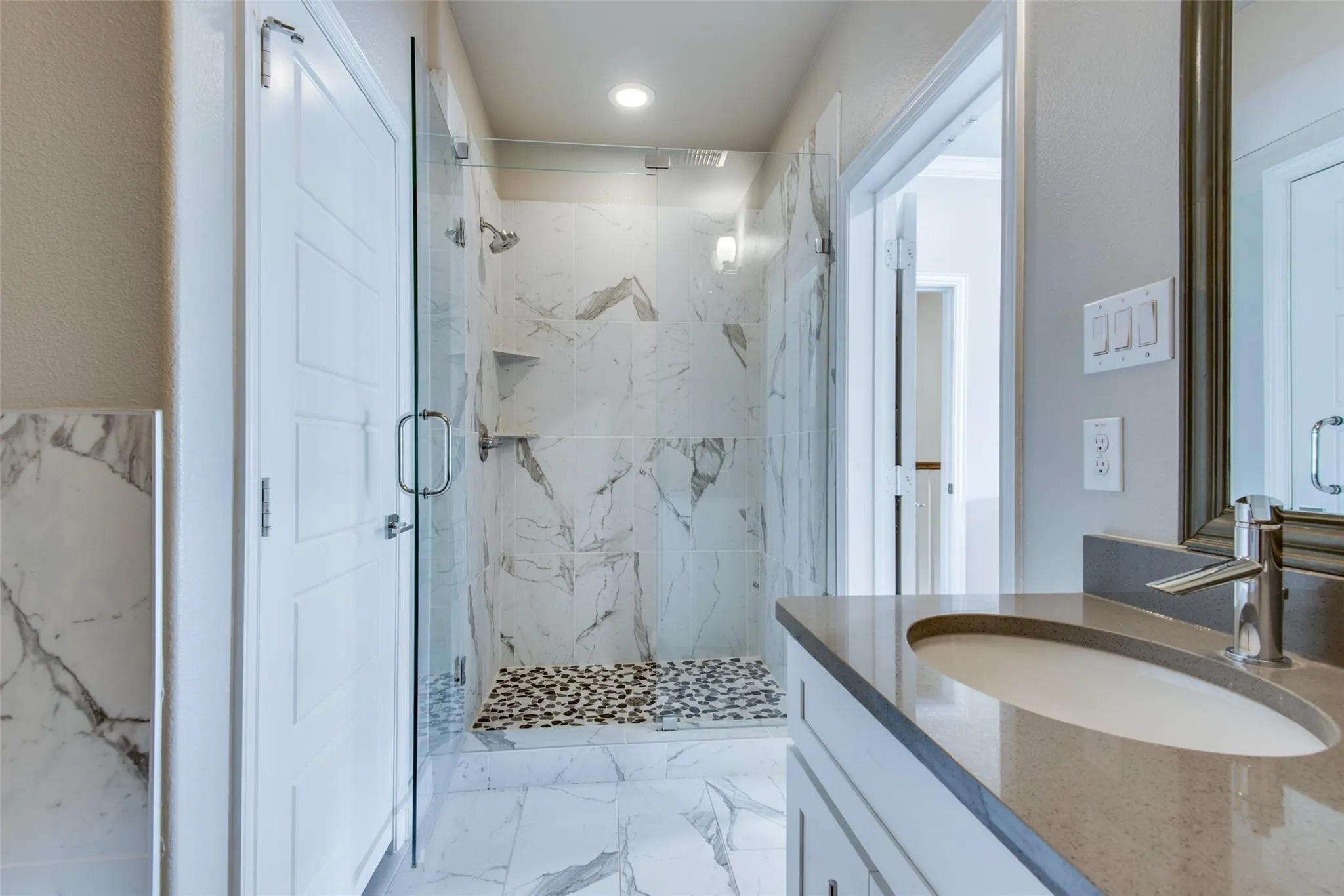 Glass-enclosed shower with marbled tile surround and pebble flooring