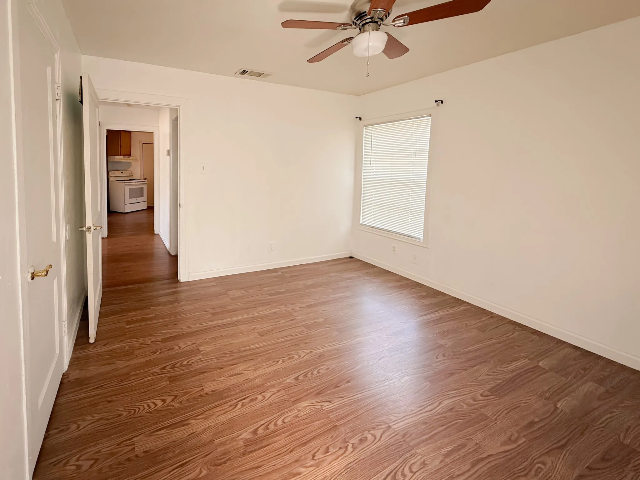 Wood-finish flooring throughout, bright white walls, window with blinds, and a ceiling fan with light fixture