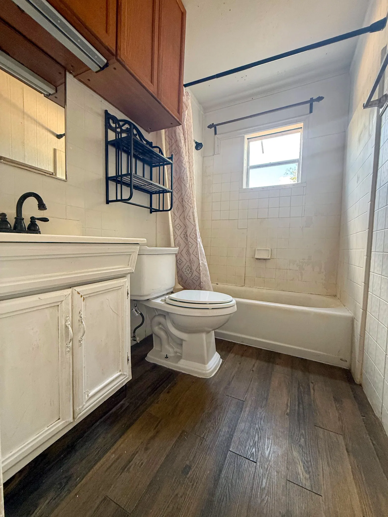 Wood-finish flooring, white vanity with cabinet storage, black faucet, built-in bathtub with shower, and tile-surround walls