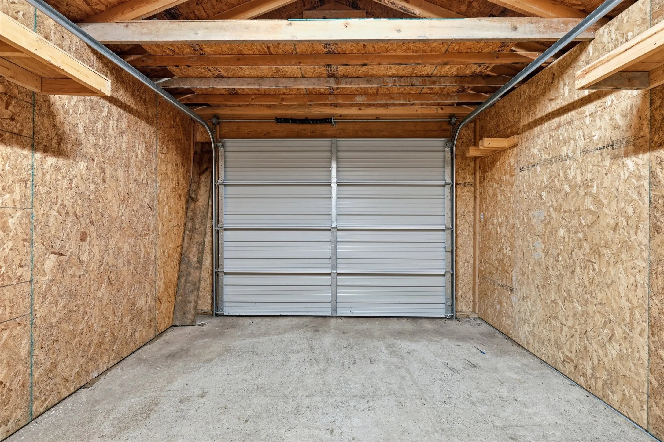 Steel roll-up garage door with a concrete floor, exposed wood ceiling beams, and oriented strand board wall finishes