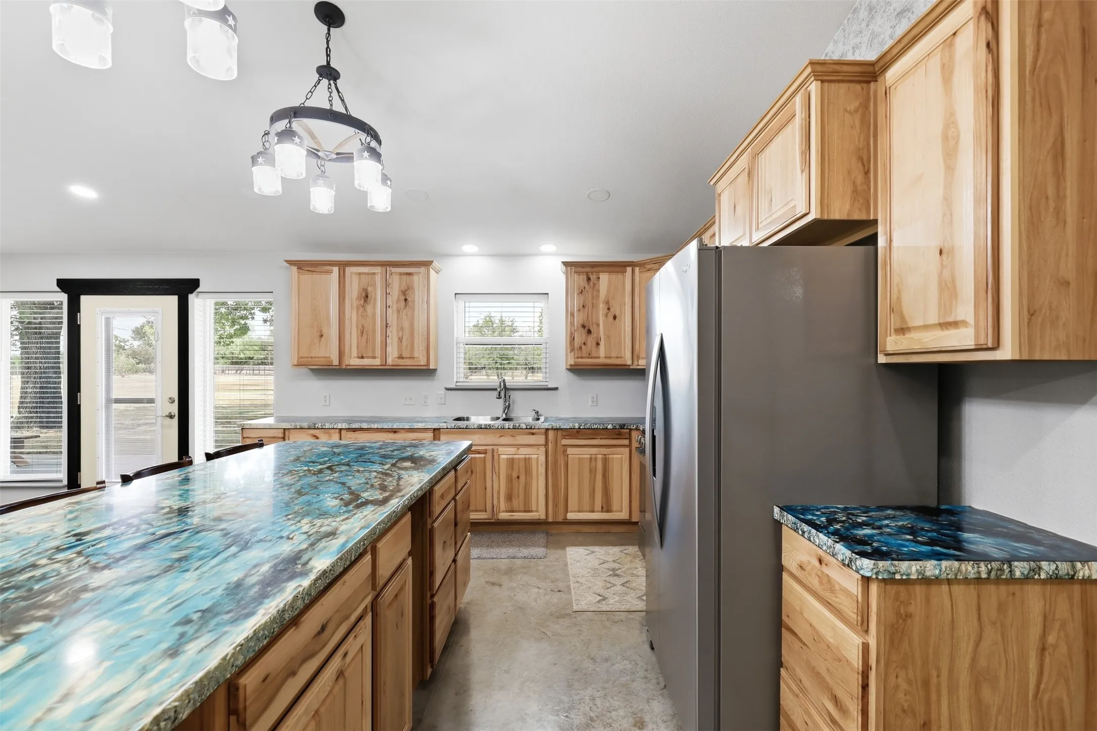 Kitchen featuring a substantial island with a patterned countertop, light wood-finish cabinetry, stainless steel refrigerator, and recessed lighting