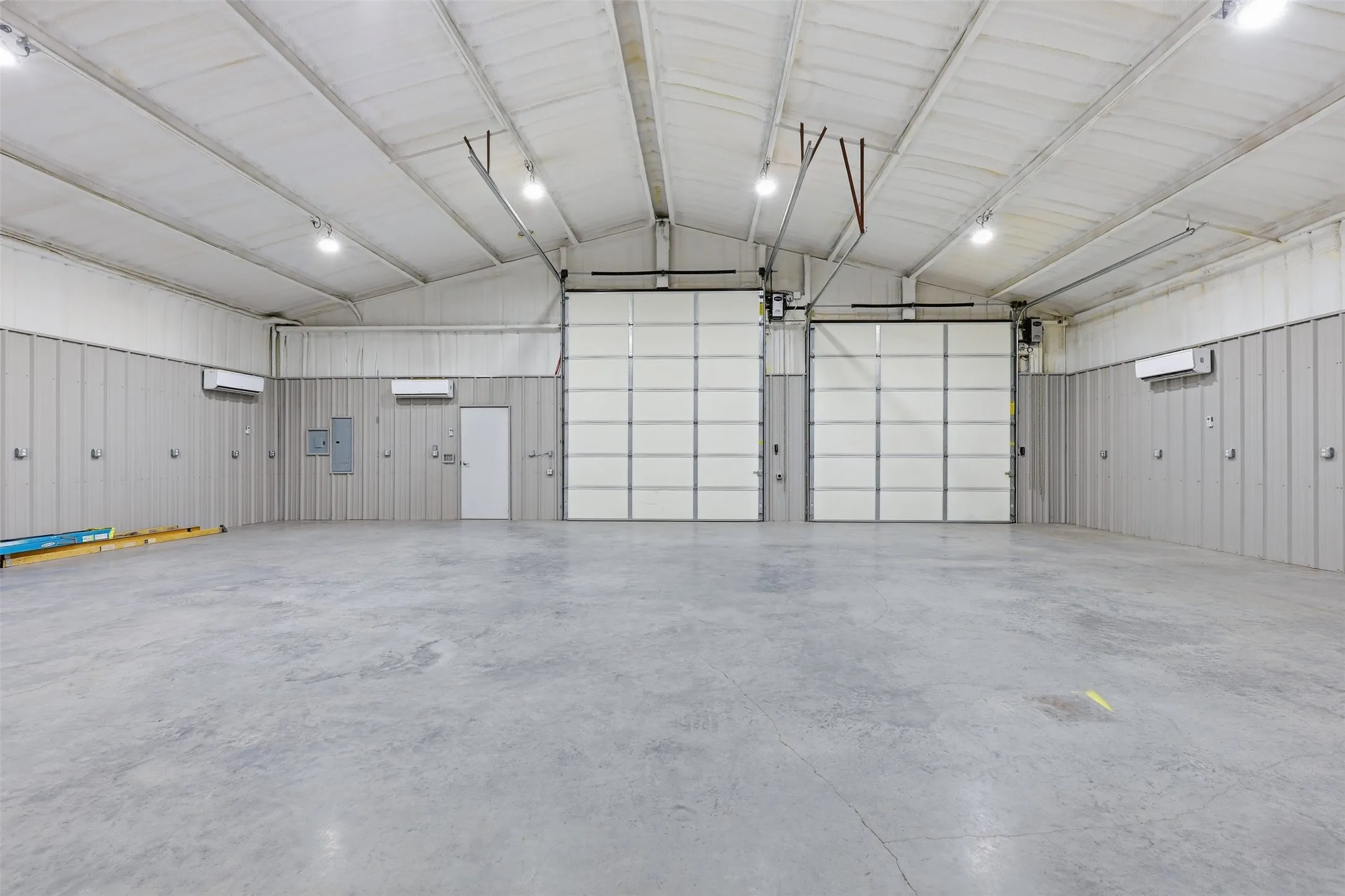 Expansive concrete flooring with high ceilings, two overhead doors, and multiple wall-mounted HVAC units