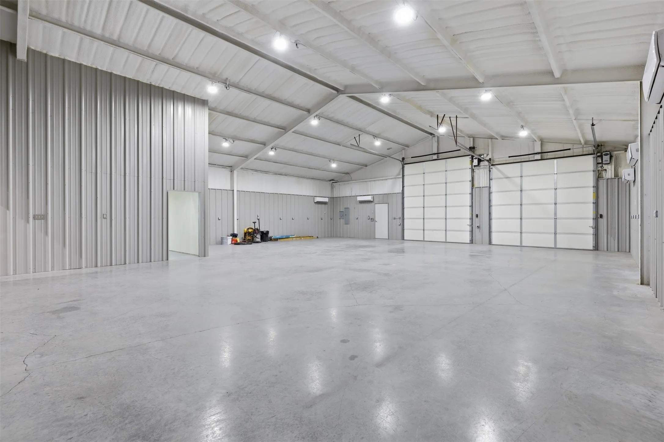 Spacious commercial interior featuring a high vaulted ceiling, two large overhead doors, multiple wall-mounted HVAC units, and polished concrete flooring