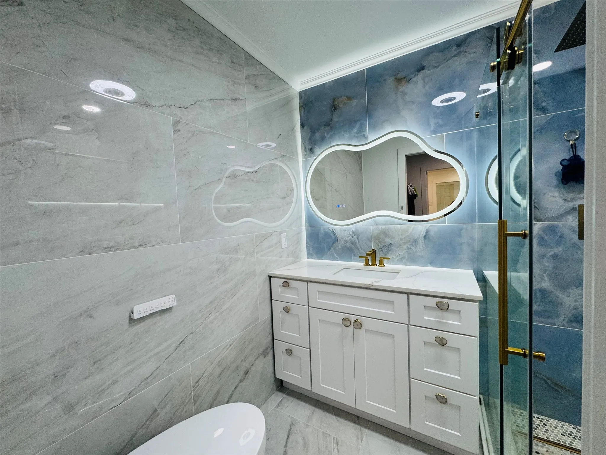 White vanity with integrated sink, gold-finish faucet, and illuminated mirror, complemented by floor-to-ceiling tile work and a glass-enclosed shower