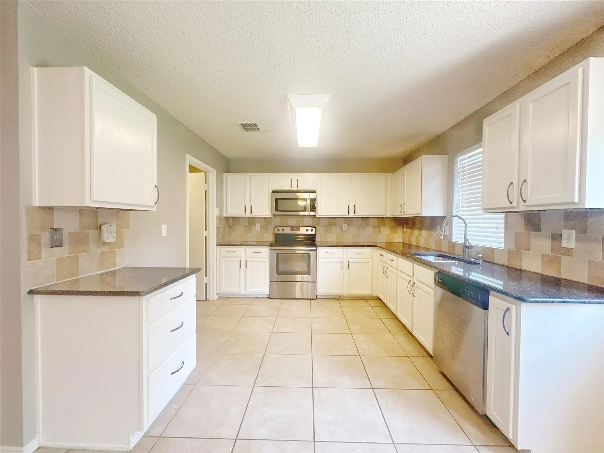 Kitchen featuring white cabinetry, stainless steel appliances, dark countertops, a tiled backsplash, and light-toned tile flooring