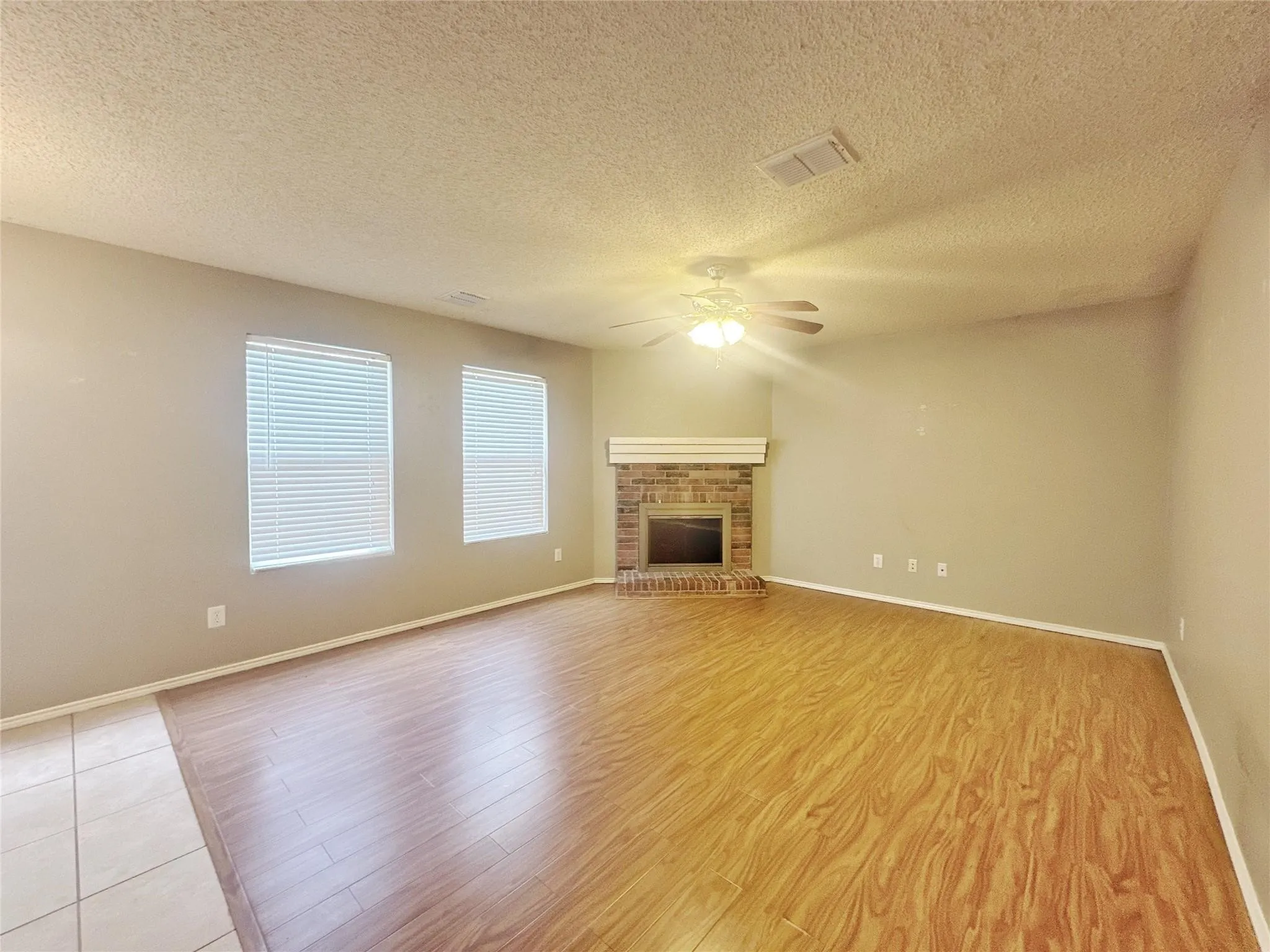 Spacious living area featuring wood-finish flooring, a brick fireplace with a white mantel, two windows with blinds, and a ceiling fan