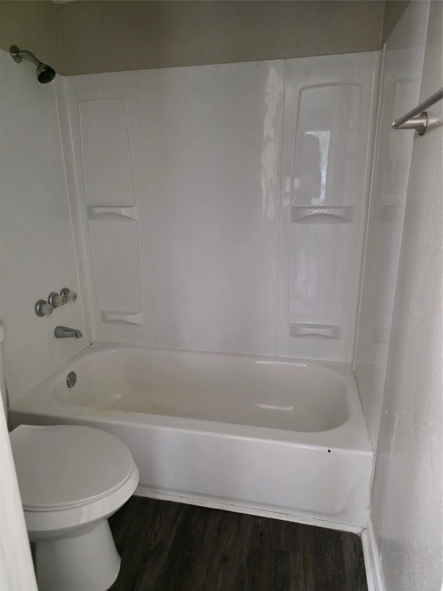 White tub and shower combination with chrome fixtures and dark wood-finish flooring