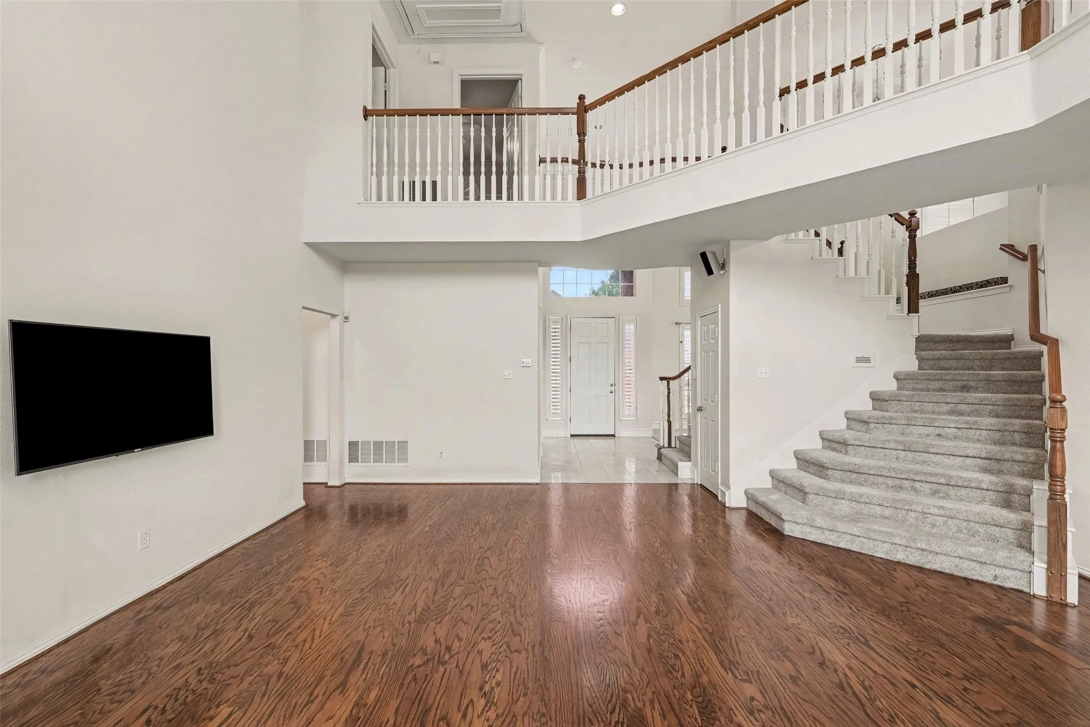 Two-story living area with wood-finish flooring, a grand carpeted staircase, and a second-story balcony featuring wood handrails
