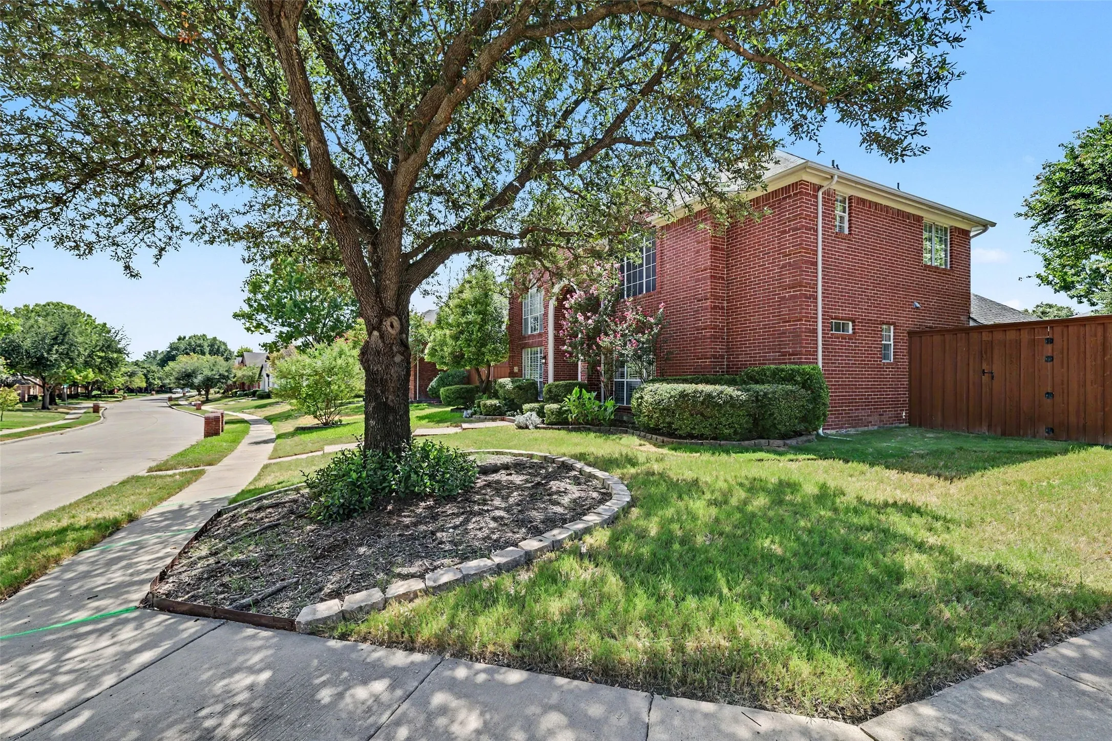 Brick exterior with multiple windows, mature front yard tree, established shrubbery, and a wood privacy fence