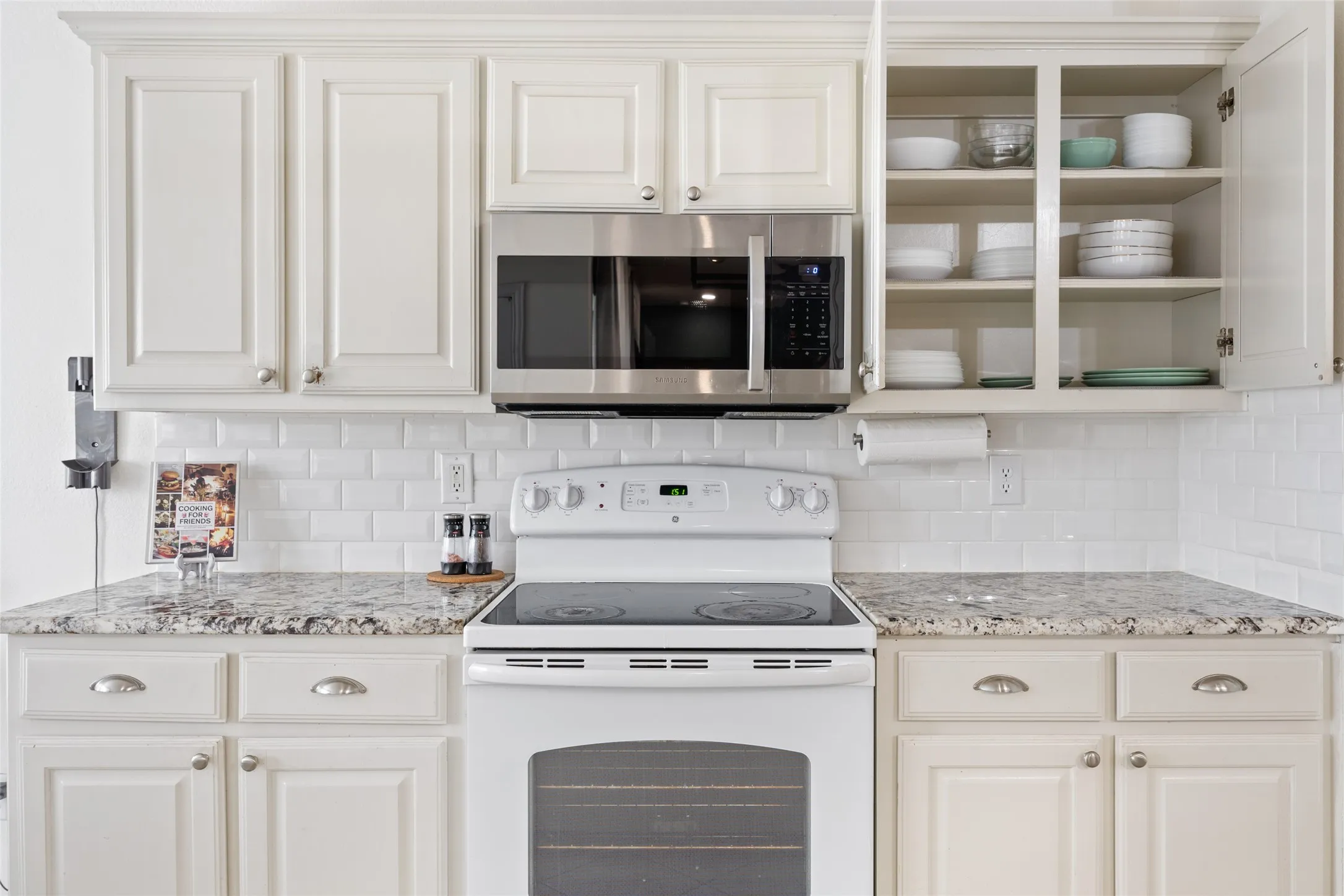 White cabinetry with brushed nickel hardware, subway tile backsplash, light-toned stone countertops, and stainless steel microwave