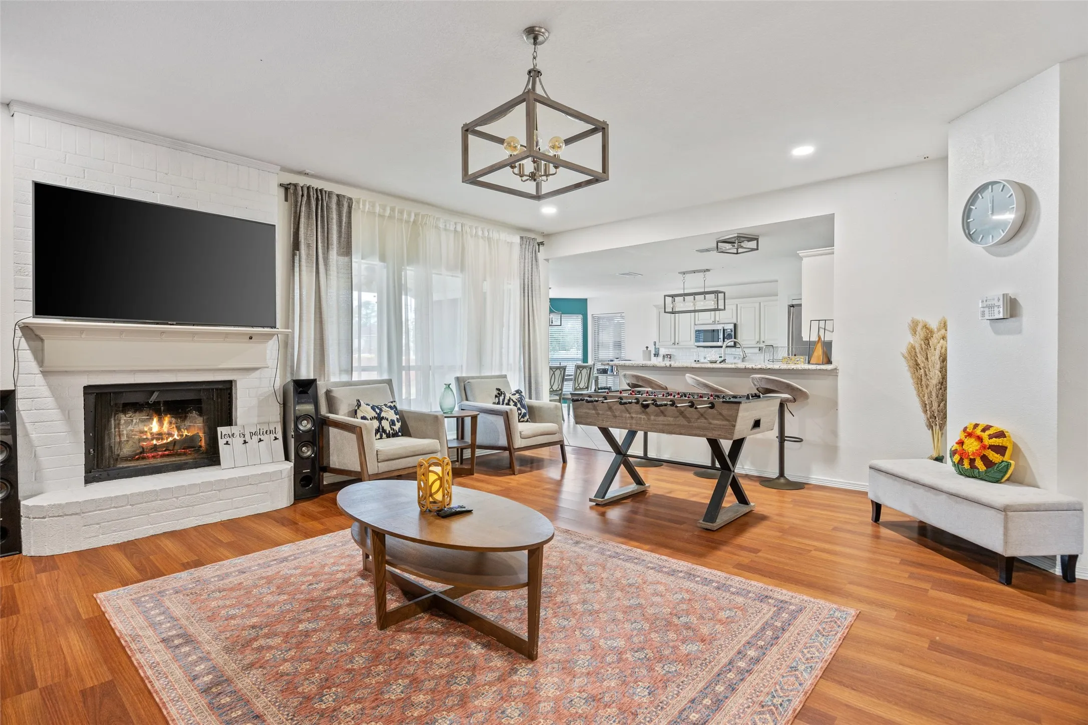 Wood-finish flooring throughout, featuring a white brick fireplace with a mantel, recessed lighting, and a geometric chandelier