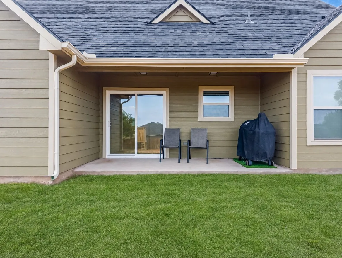 Covered concrete patio with sliding glass door access, flanked by horizontal siding and a shingled roof