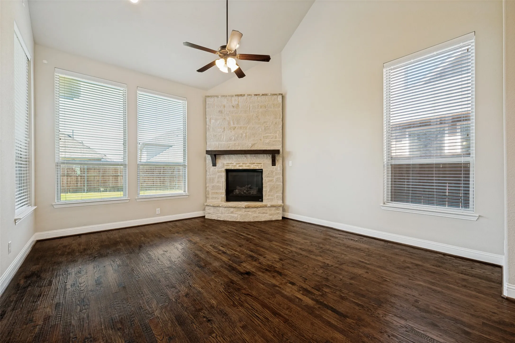 Living area featuring dark wood-finish flooring, a corner stone fireplace with a dark wood mantel, and a vaulted ceiling with a ceiling fan