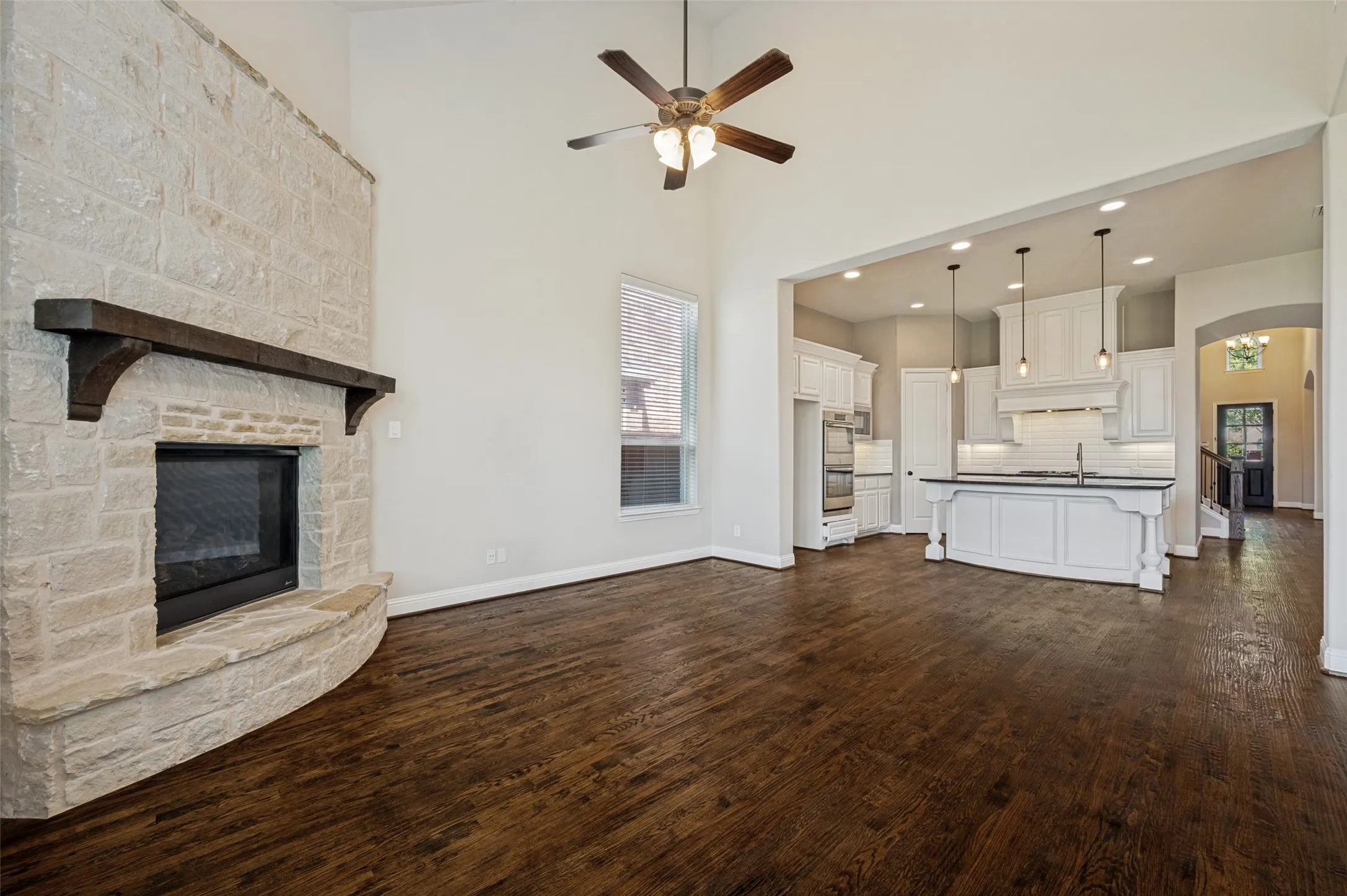 Living area featuring a stone-clad fireplace with a dark wood mantel, high ceilings, wood-finish flooring, and an open concept layout to the kitchen