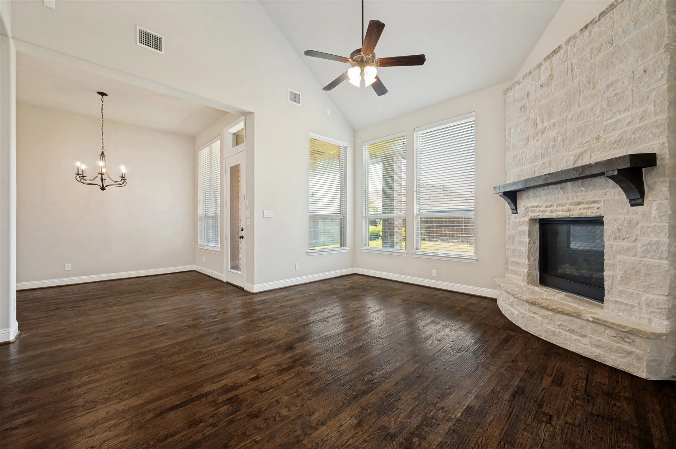Spacious living area featuring high vaulted ceilings, expansive wood-finish flooring, a stone-clad fireplace with a dark wood mantel, a ceiling fan, and multiple windows
