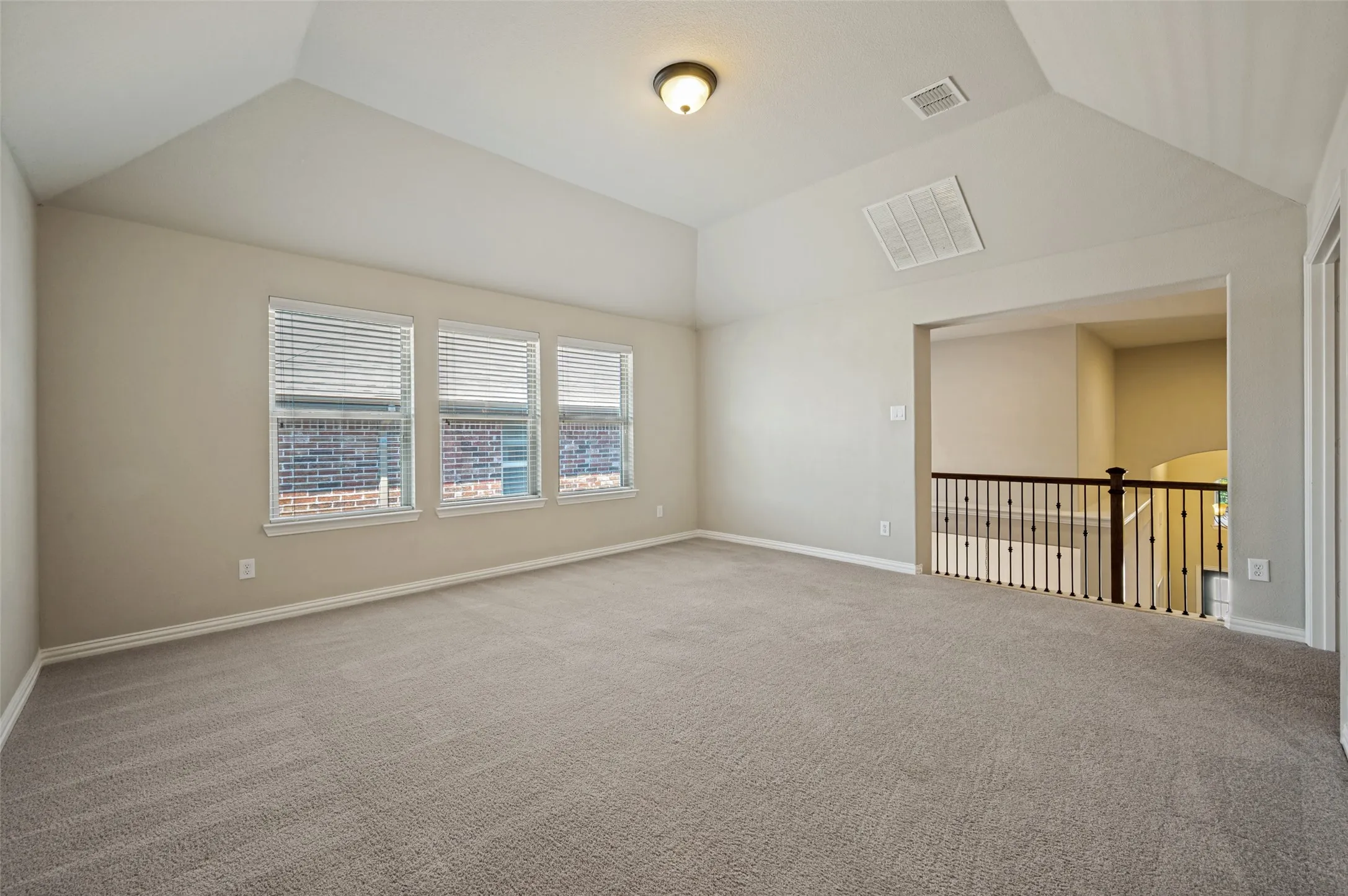 Spacious room featuring vaulted ceilings, light-colored carpeting, three windows with horizontal blinds, a ceiling-mounted light fixture, and an open railing overlooking a stairway
