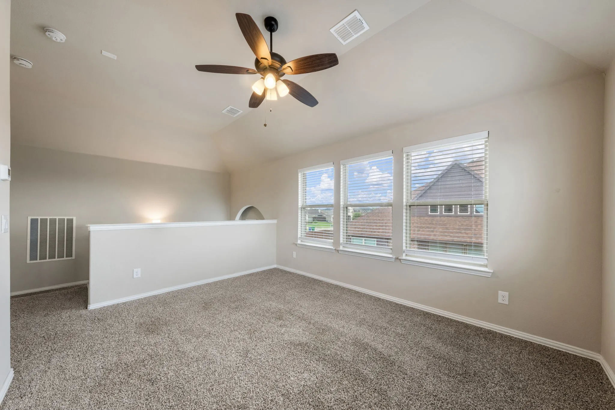 Expansive carpeted room featuring a ceiling fan with light fixture, three windows with blinds, and a partial wall with an arched opening