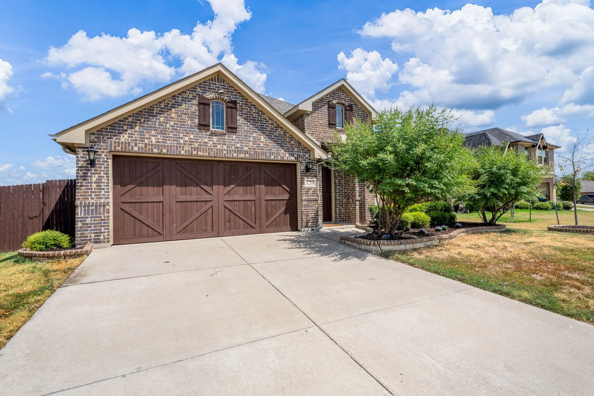 Brick facade with decorative shutters, two-car garage with wood-look doors, concrete driveway, and front landscaping with established plantings