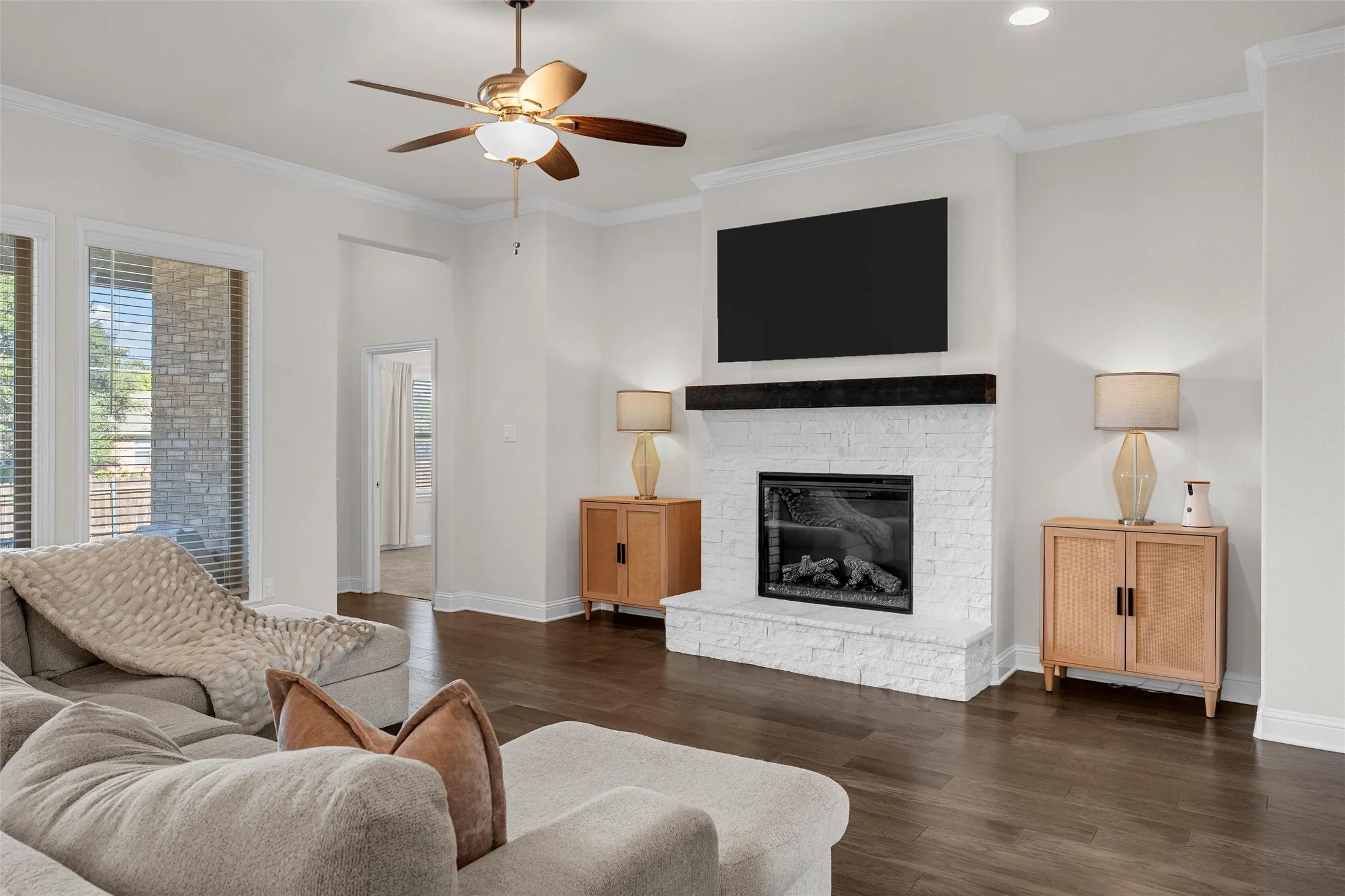 Wood-finish flooring, white brick fireplace with a dark wood mantel, ceiling fan with integrated lighting, recessed lighting, and crown molding