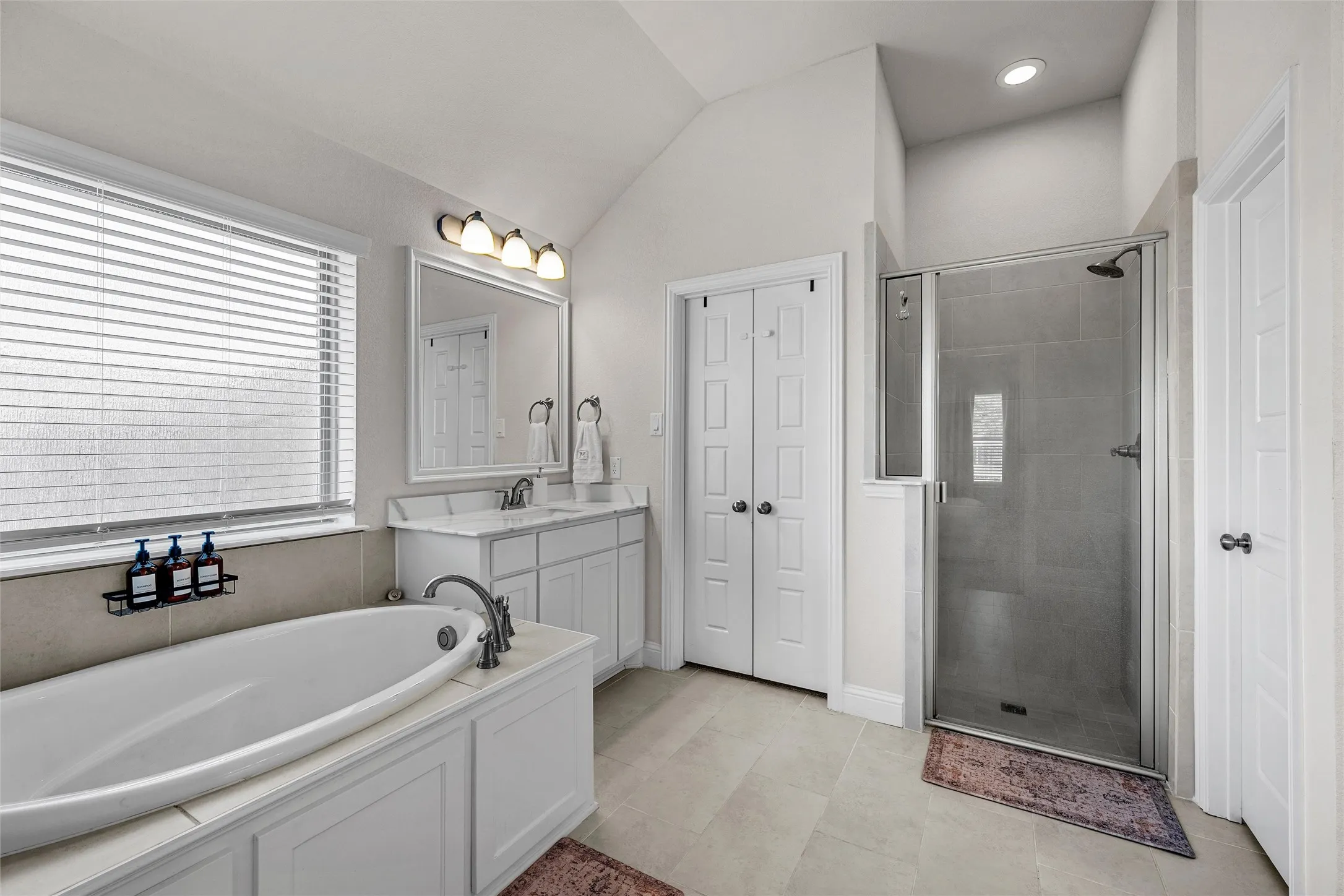 Soaking tub with tile surround, dual vanity with undermount sinks, separate glass-enclosed shower, vaulted ceiling, and neutral tile flooring