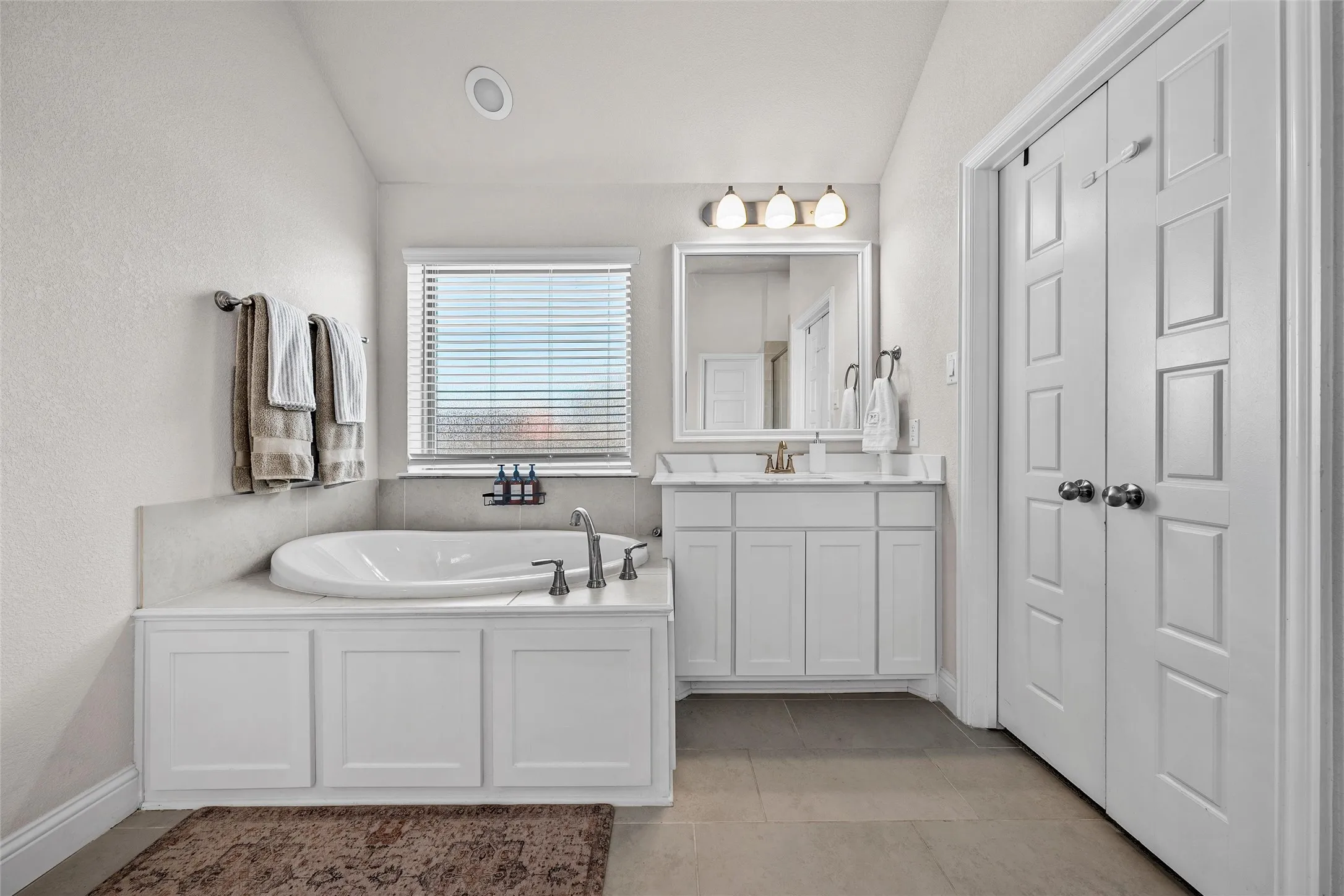 Built-in soaking tub with gray-toned tile surround, single vanity with white cabinetry and framed mirror, window with blinds, and white panel doors