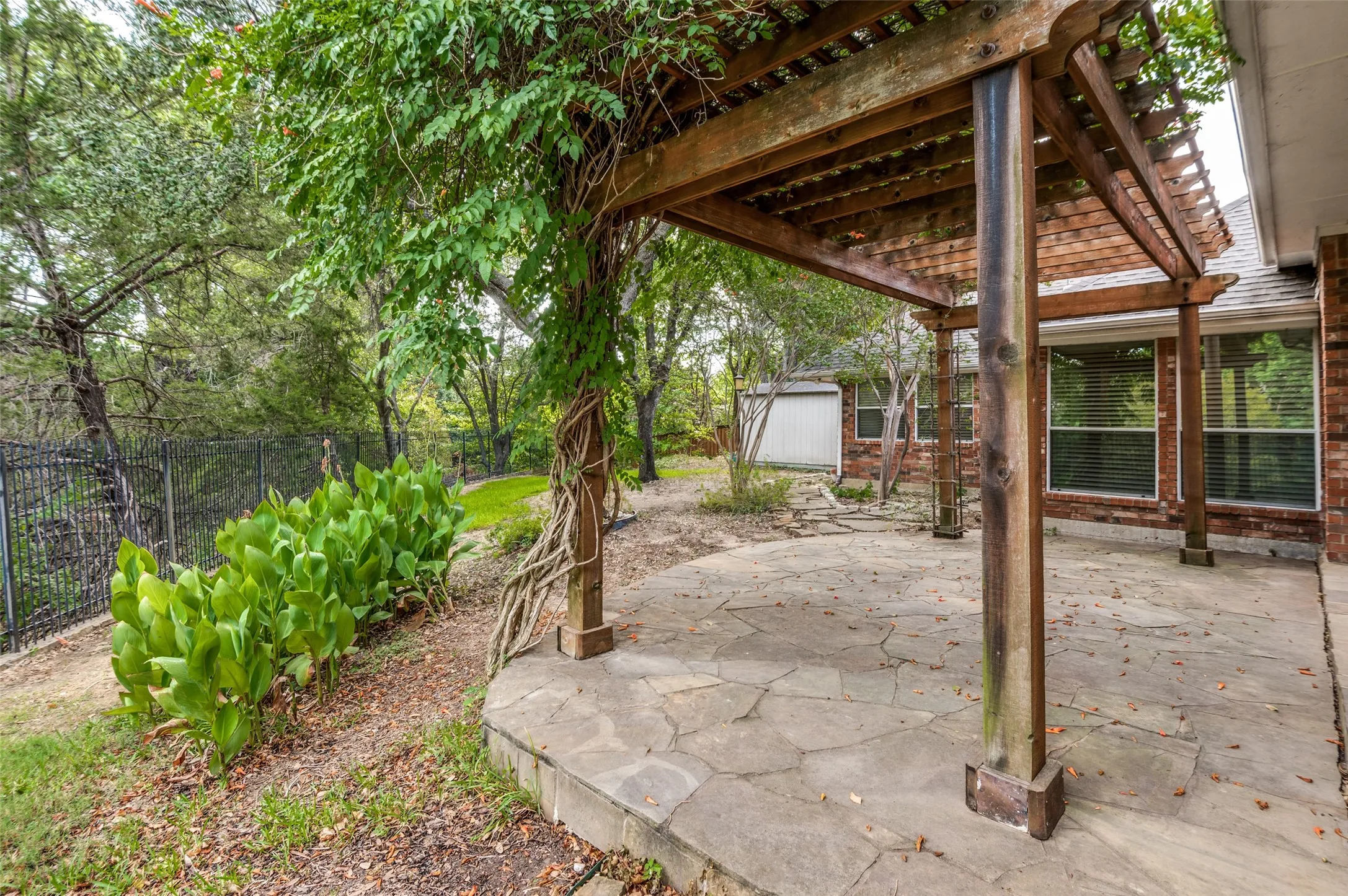 Expansive stone patio with a wood pergola, lush perimeter landscaping, and a fenced boundary
