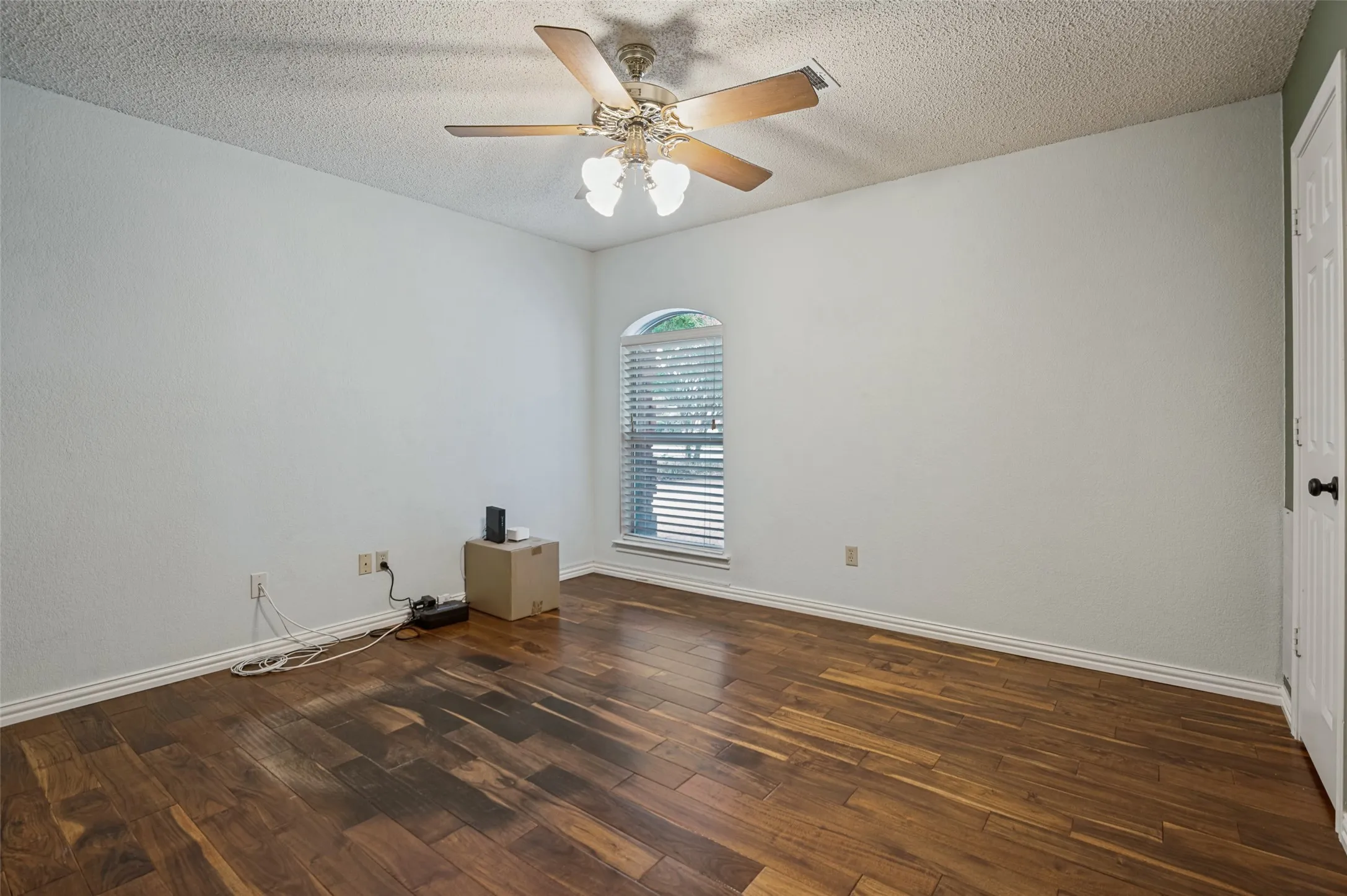 Wood-finish flooring, ceiling fan with integrated lighting, arched window with horizontal blinds, and a white paneled door