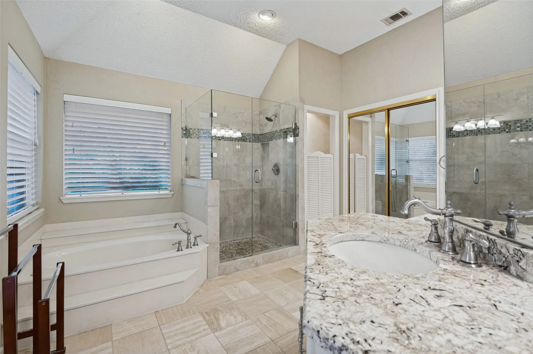 Spacious bathroom featuring a built-in soaking tub, glass-enclosed shower, large vanity with stone-finish countertop, and mirrored sliding closet doors