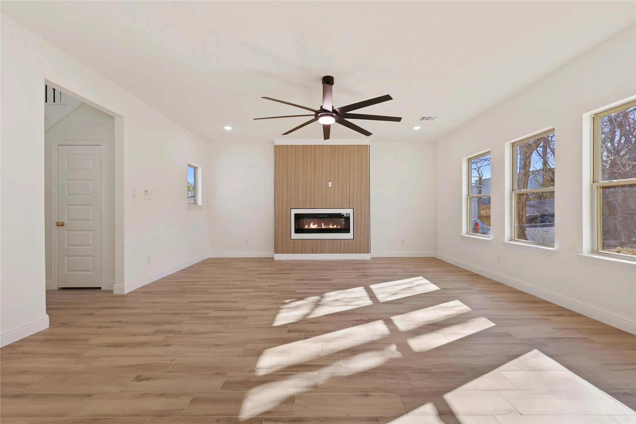 Spacious room with wood-finish flooring, an integrated electric fireplace with wood paneling, a contemporary ceiling fan, recessed lighting, and multiple windows