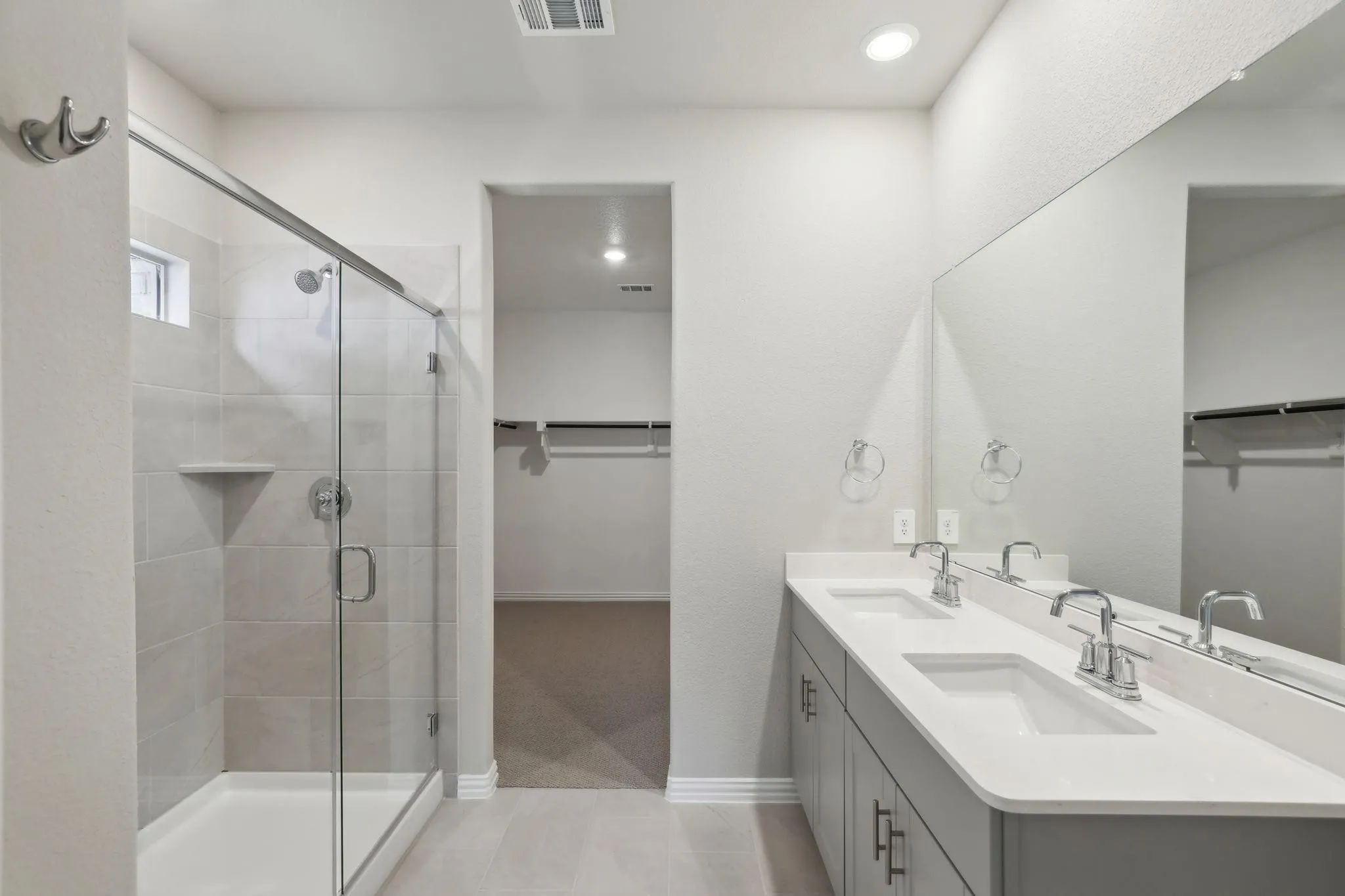 Dual vanity with undermount sinks and chrome fixtures, frameless mirror, tiled shower with glass enclosure, recessed lighting, and an adjacent walk-in closet with built-in shelving