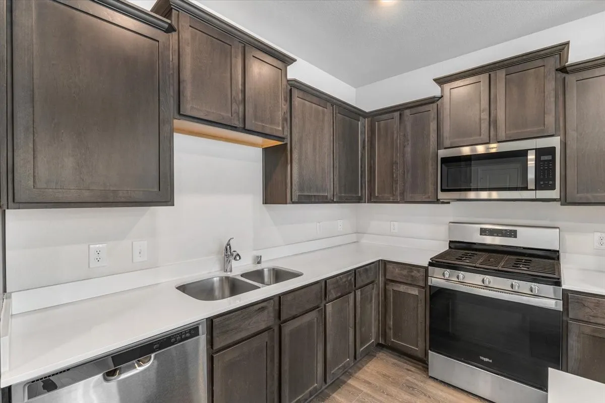 Kitchen featuring dark wood-finish cabinetry, light countertops, a stainless steel double basin sink, a stainless steel dishwasher, and a stainless steel range with an overhead microwave