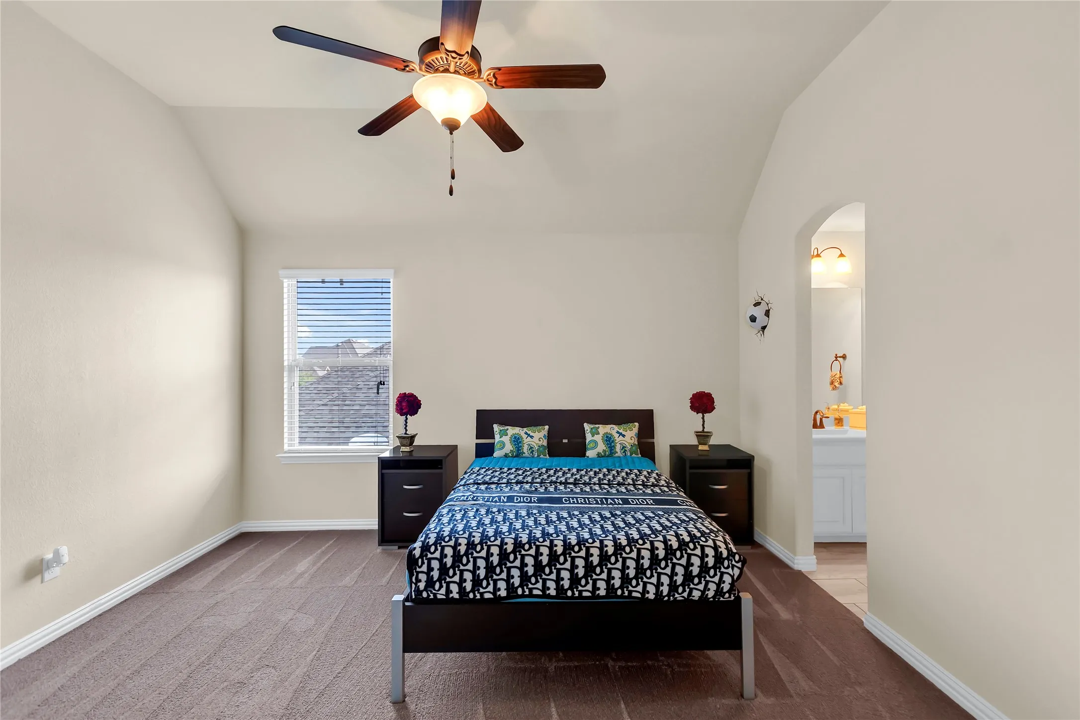 Vaulted ceiling with a ceiling fan, window with blinds, carpeted flooring, and an arched doorway leading to a bathroom with a white vanity and gold-tone fixtures