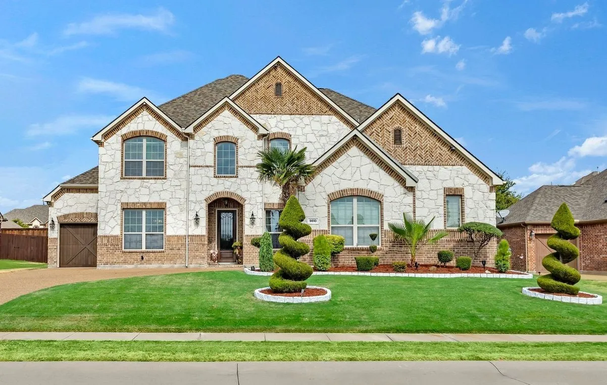 Two-story exterior featuring stone and brick accents, a gabled roofline, arched windows, a wood-finish garage door, and a landscaped front yard with decorative plantings