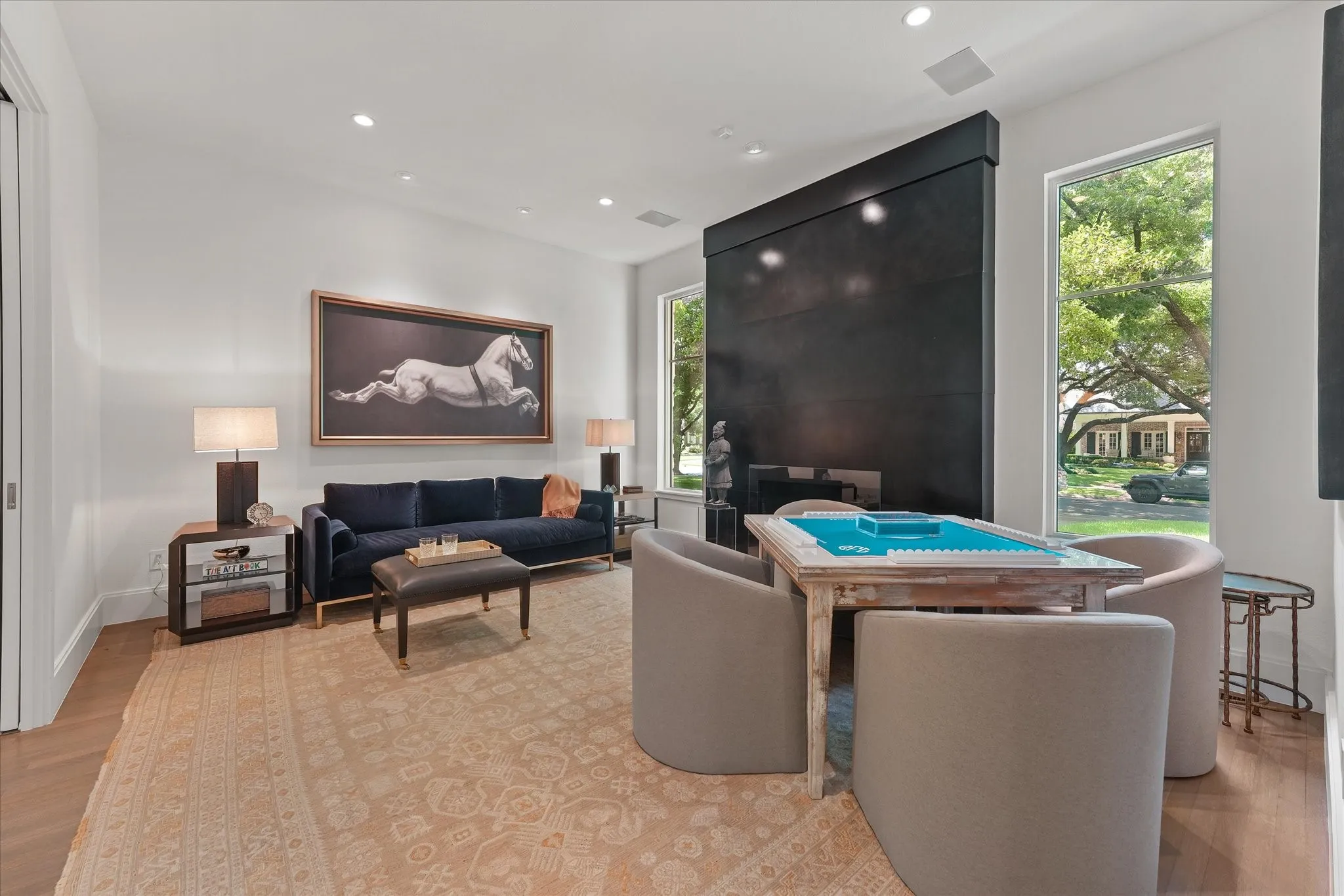 A tall, dark fireplace surround contrasts with white walls, while windows on either side overlook the front yard.