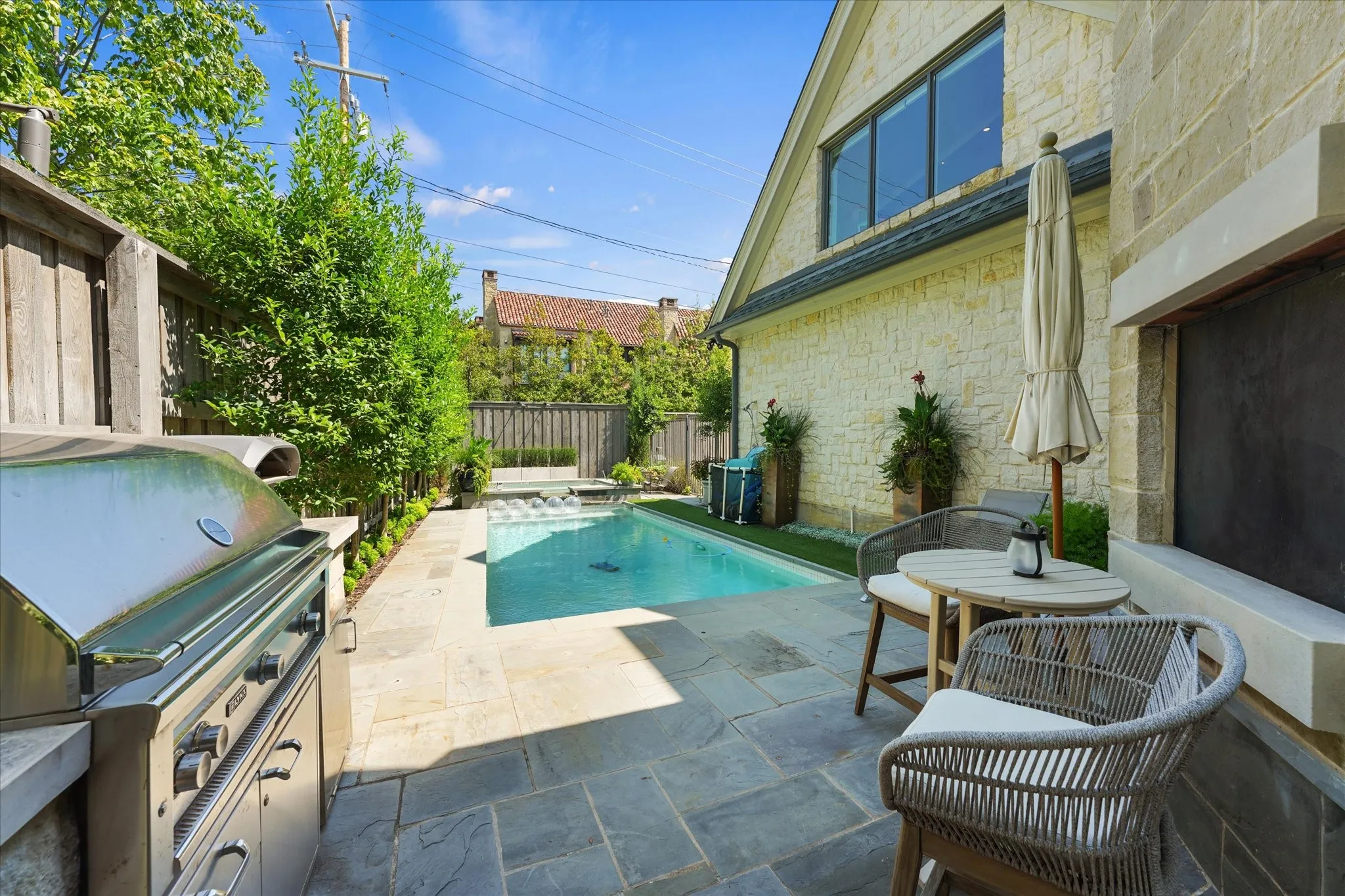 A built-in stainless steel grill lines the stone patio beside the rectangular pool, with trees and a wood fence along the perimeter.