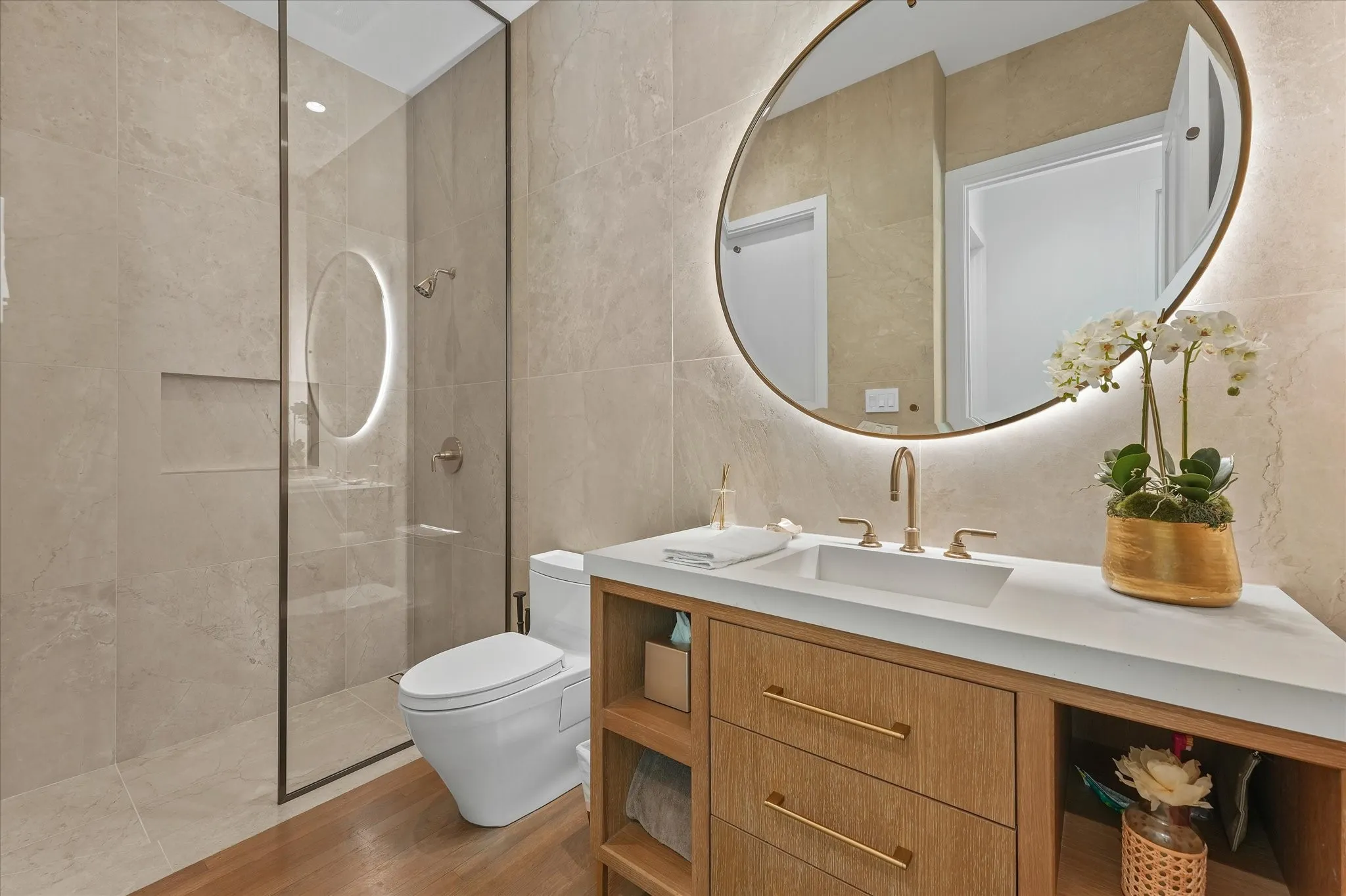 A backlit round mirror hangs above a wood vanity beside a glass-enclosed shower lined with large-format tile.