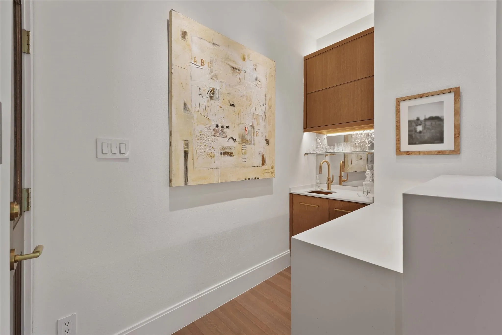 Wood cabinets frame a wet bar and ice machine with a mirrored backsplash, glass shelving, white countertops and a gold-toned faucet.