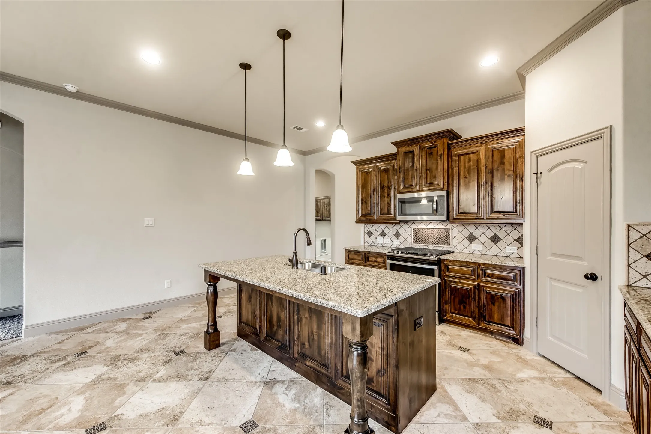 Kitchen featuring a center island with a built-in sink, pendant lighting, stone tile flooring, stained wood cabinetry, and stainless steel appliances