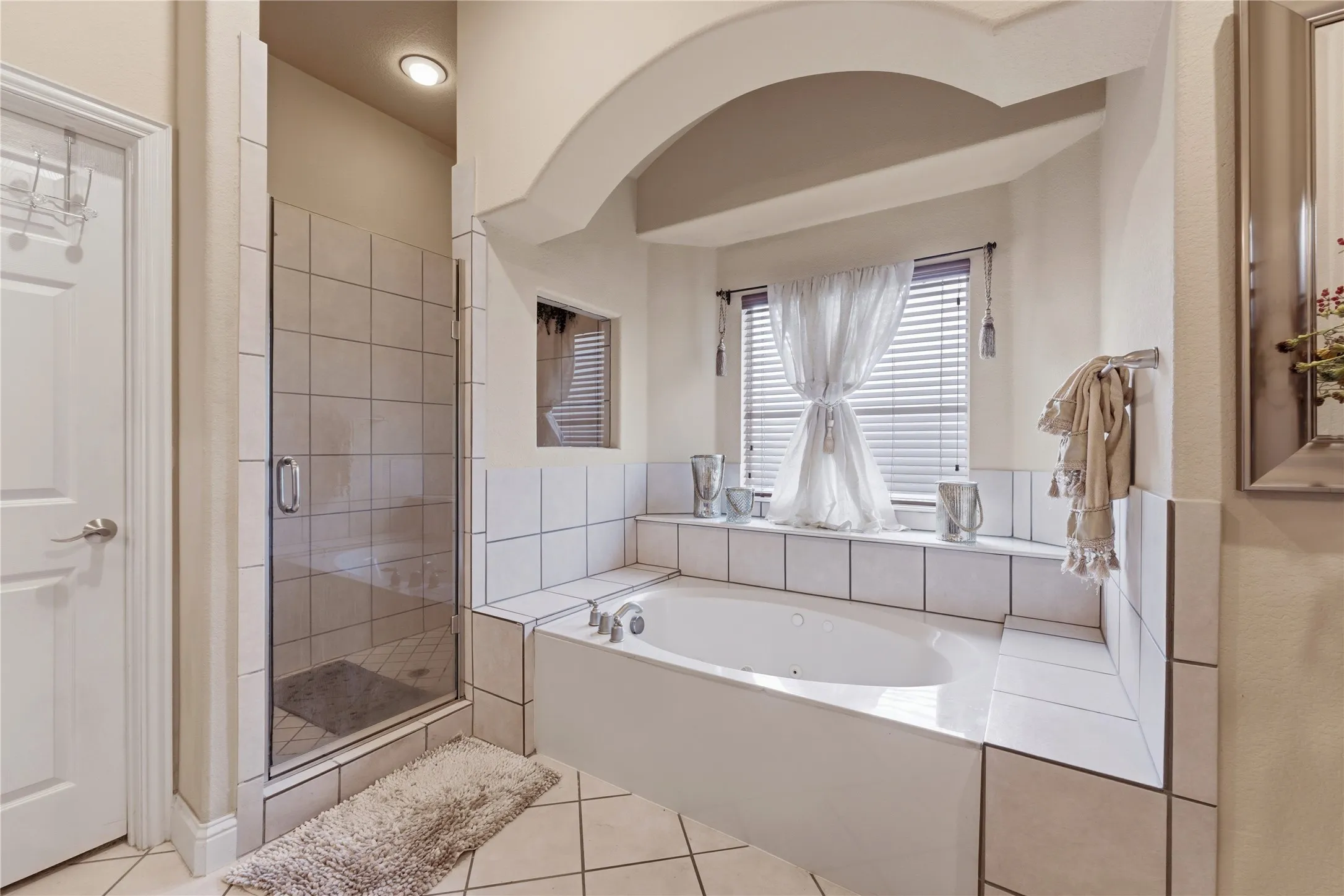 Soaking tub set within a tiled surround, arched alcove, separate glass-enclosed shower, tiled flooring, and window with blinds