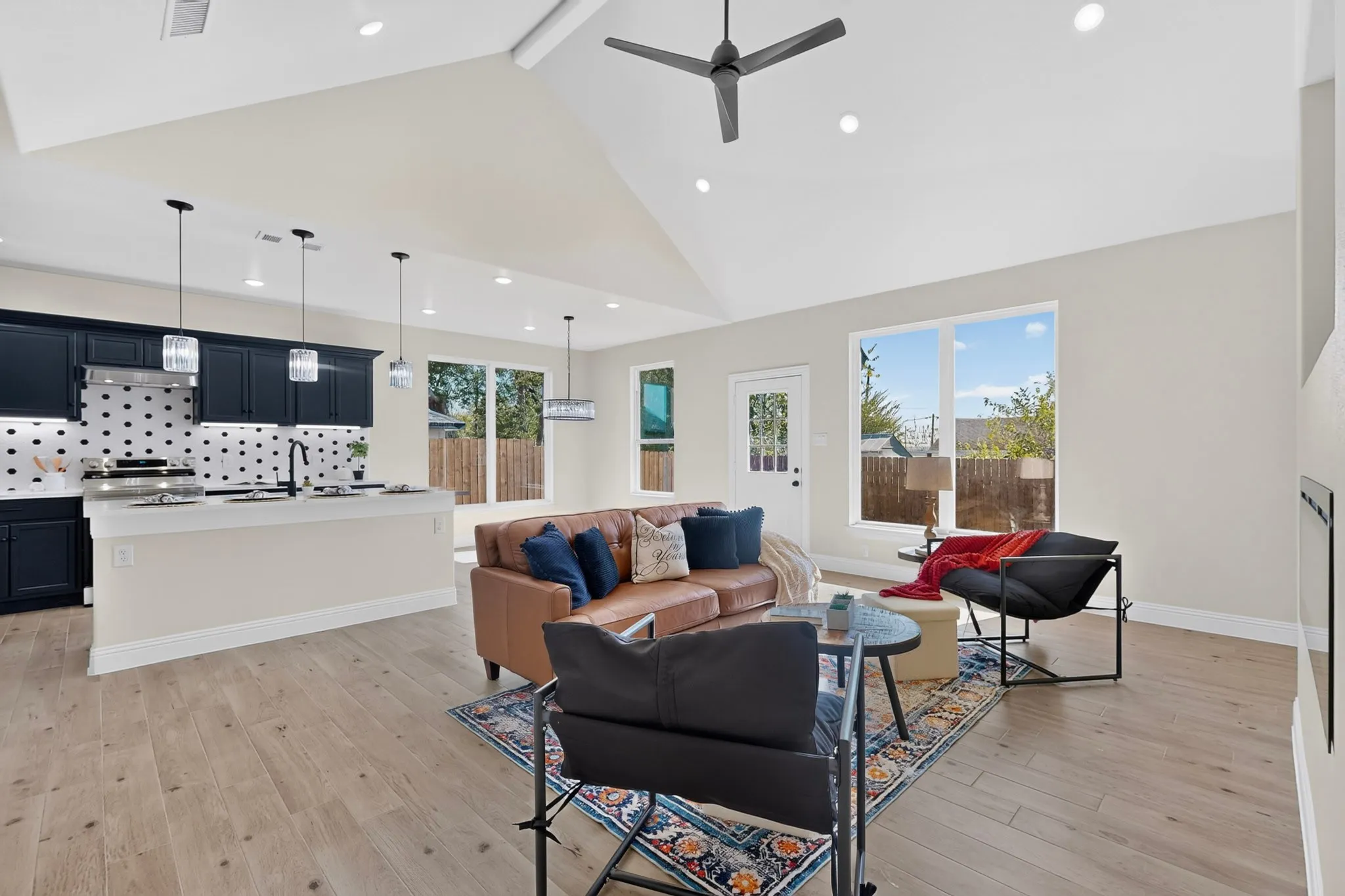 Vaulted ceiling with recessed lighting and ceiling fan, wood-finish flooring, dark cabinetry with light countertops and patterned backsplash, kitchen island with pendant lighting, and multiple windows
