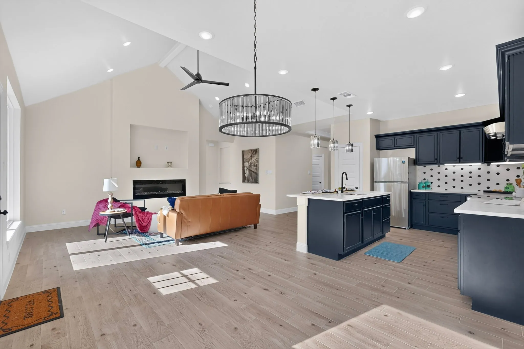 Vaulted ceilings with recessed lighting, linear fireplace with built-in niche, wood-finish flooring, kitchen island with sink and pendant lighting, and dark-toned cabinetry with white countertops
