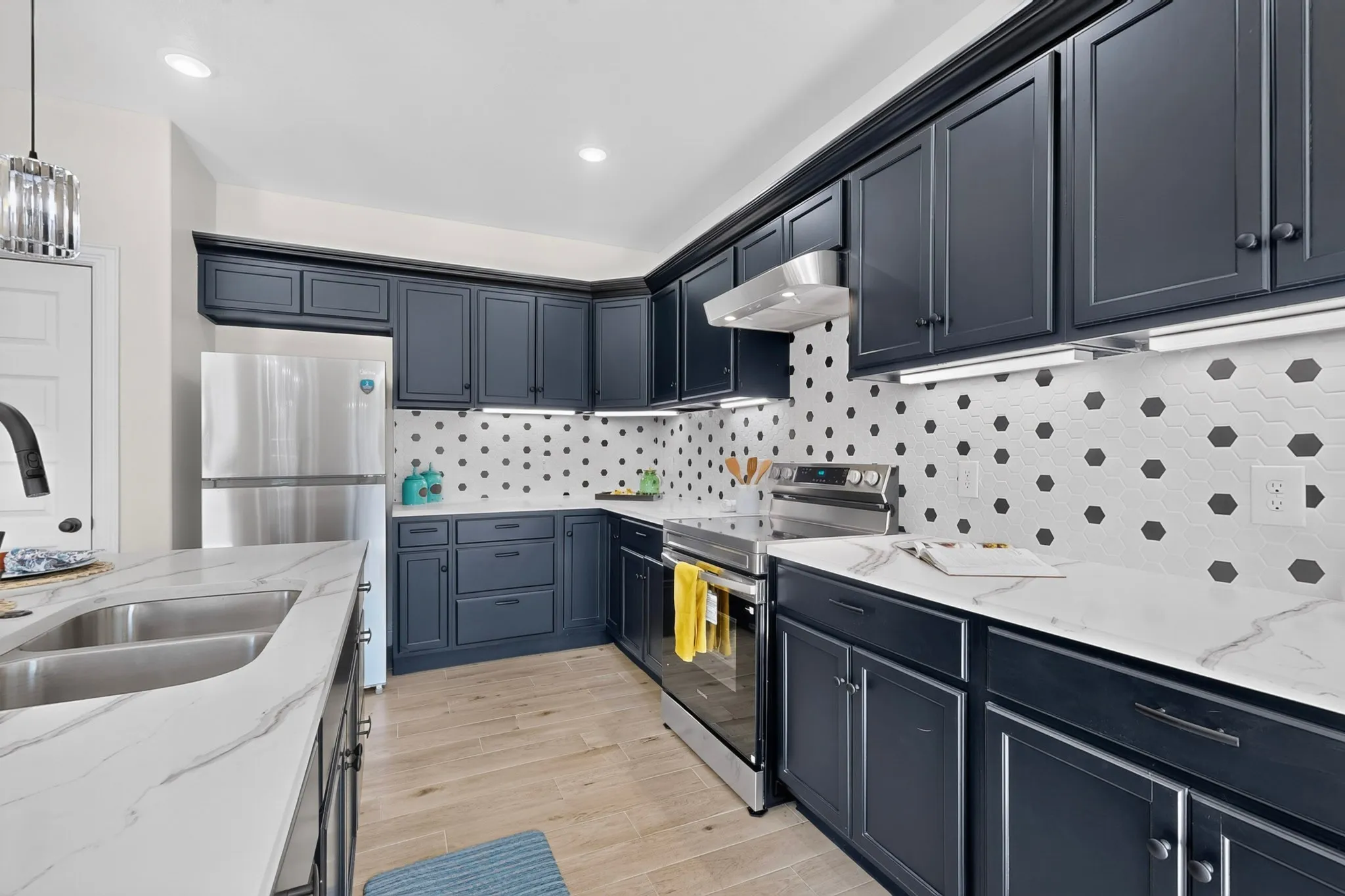 Kitchen featuring dark blue cabinetry, light-toned countertops with veining, a hexagon-patterned backsplash, stainless steel appliances, and light wood-finish flooring