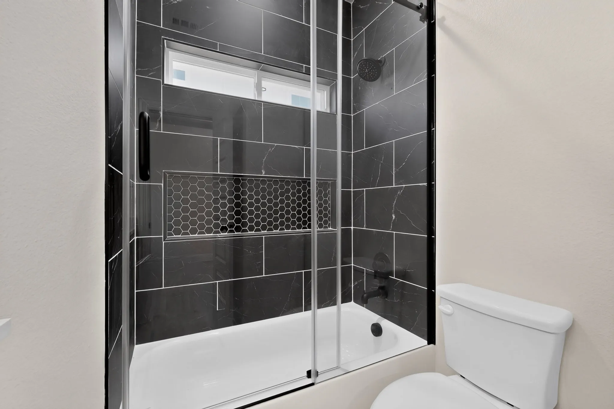 Bathtub with black-tiled surround, glass enclosure, recessed hexagon tile niche, and matte black shower fixtures