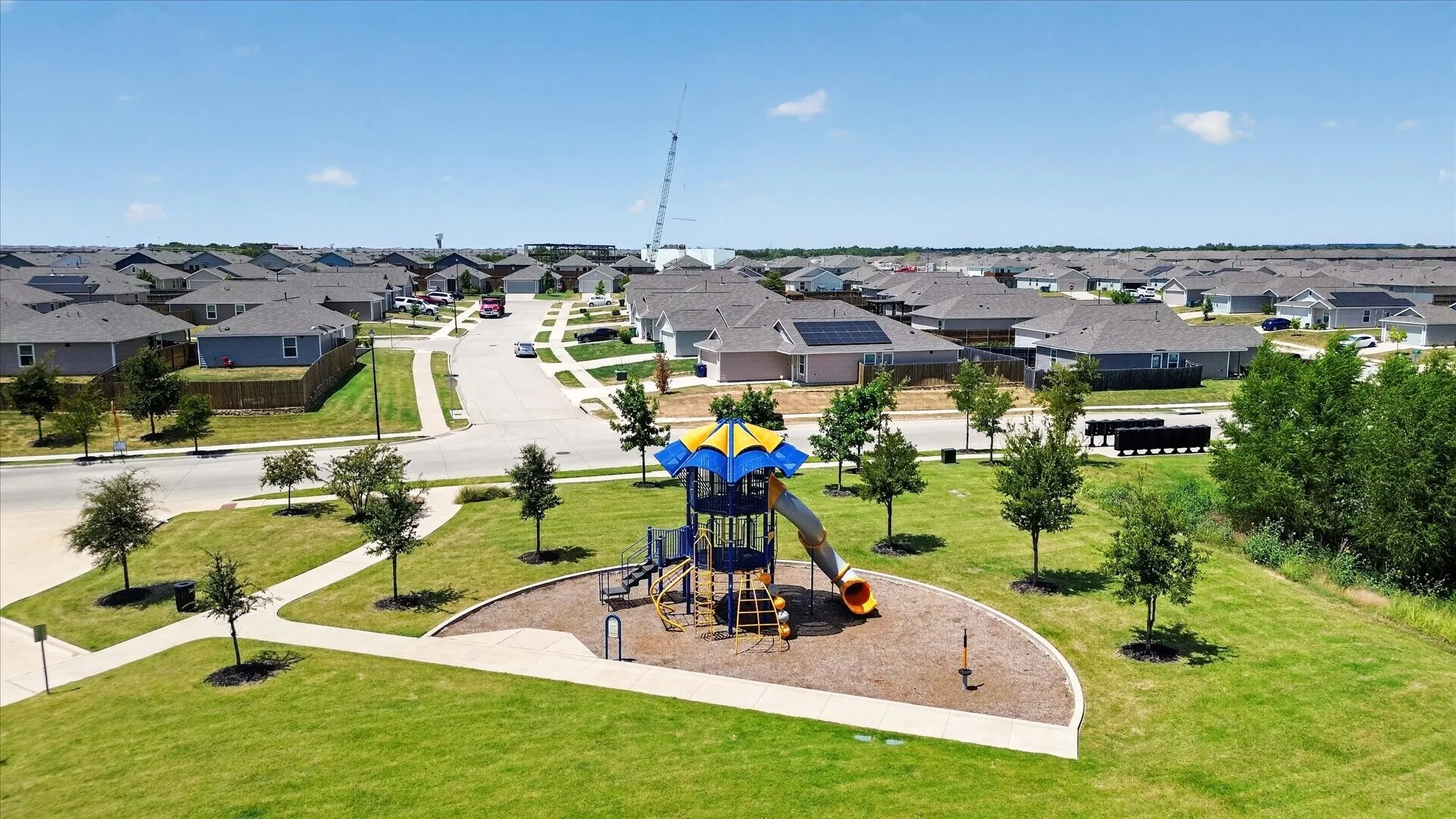 Community green space featuring a playground with slides and climbing elements, surrounded by a manicured lawn, paved walkways, and new construction homes